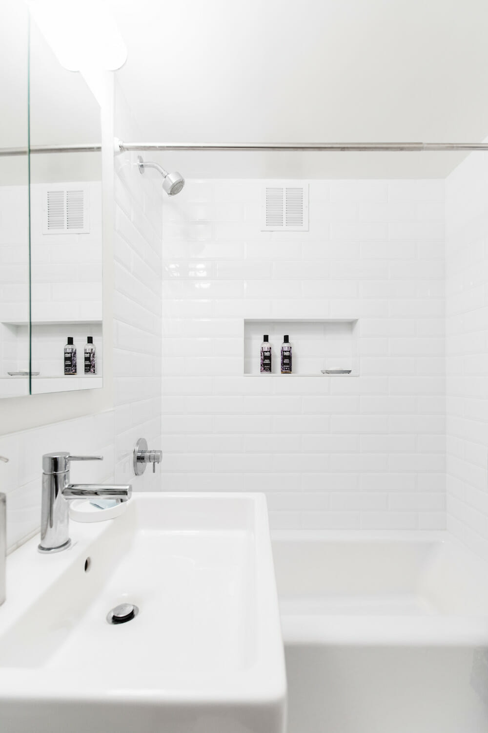 all white bathroom with white tiles on shower walls and recessed shelf and bathtub and farmhouse sink with nickel faucet and fixtures after renovation