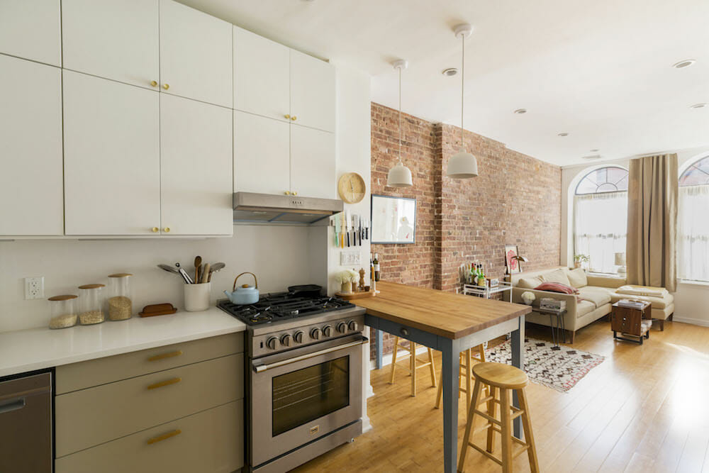 white overhead kitchen cabinets and white countertop and beige under counter cabinets and stainless steel appliances and peninsula island with butcher block countertop and exposed brick wall and hardwood floors after renovation