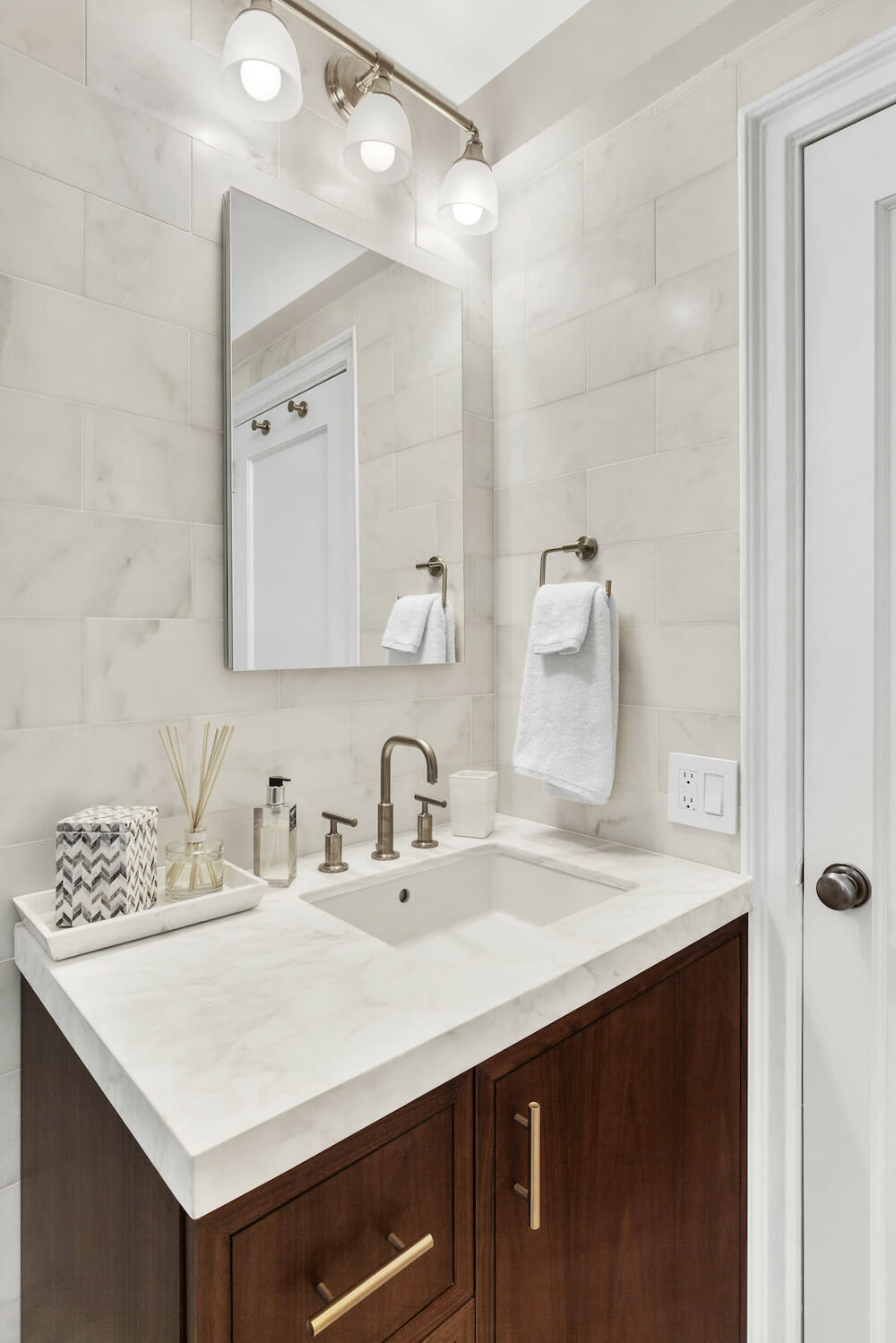 white marble tiles on bathroom wall and white marble countertop with undermount sink and brass faucet and wall mounted lights above medicine cabinet with mirror after renovation