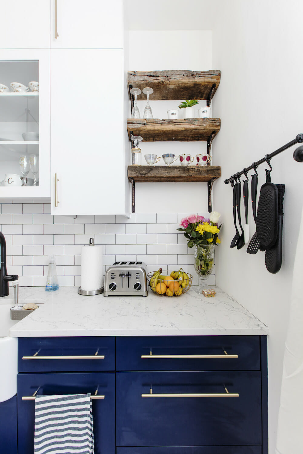 kitchen with white overhead cabinets and white countertop and navy blue cabinets under counter and floating wooden shelves and backsplash with white subway tiles after renovation