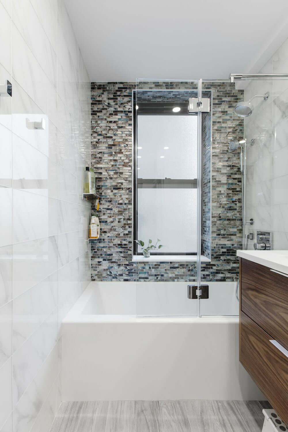 bathtub with mosaic tiles on one wall and marble wall tiles and glass doorand gray floor tiles and floating vanity after renovation