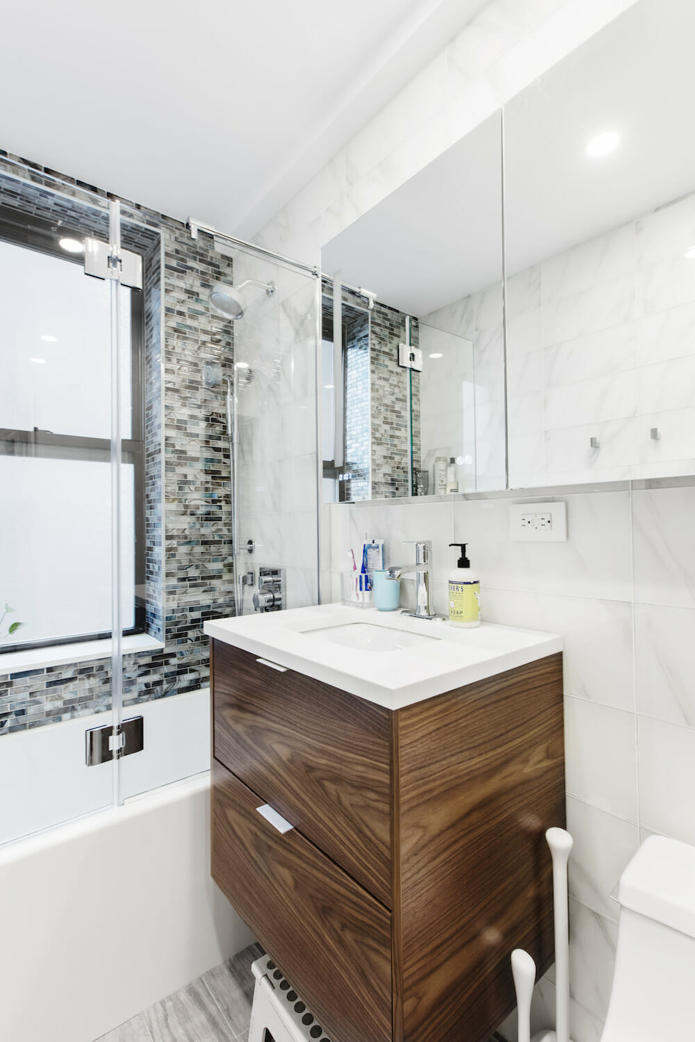 floating wooden bathroom vanity with white countertop and mirror and white tiles on walls and bathtub with mosaic wall tiles and glass door after renovation