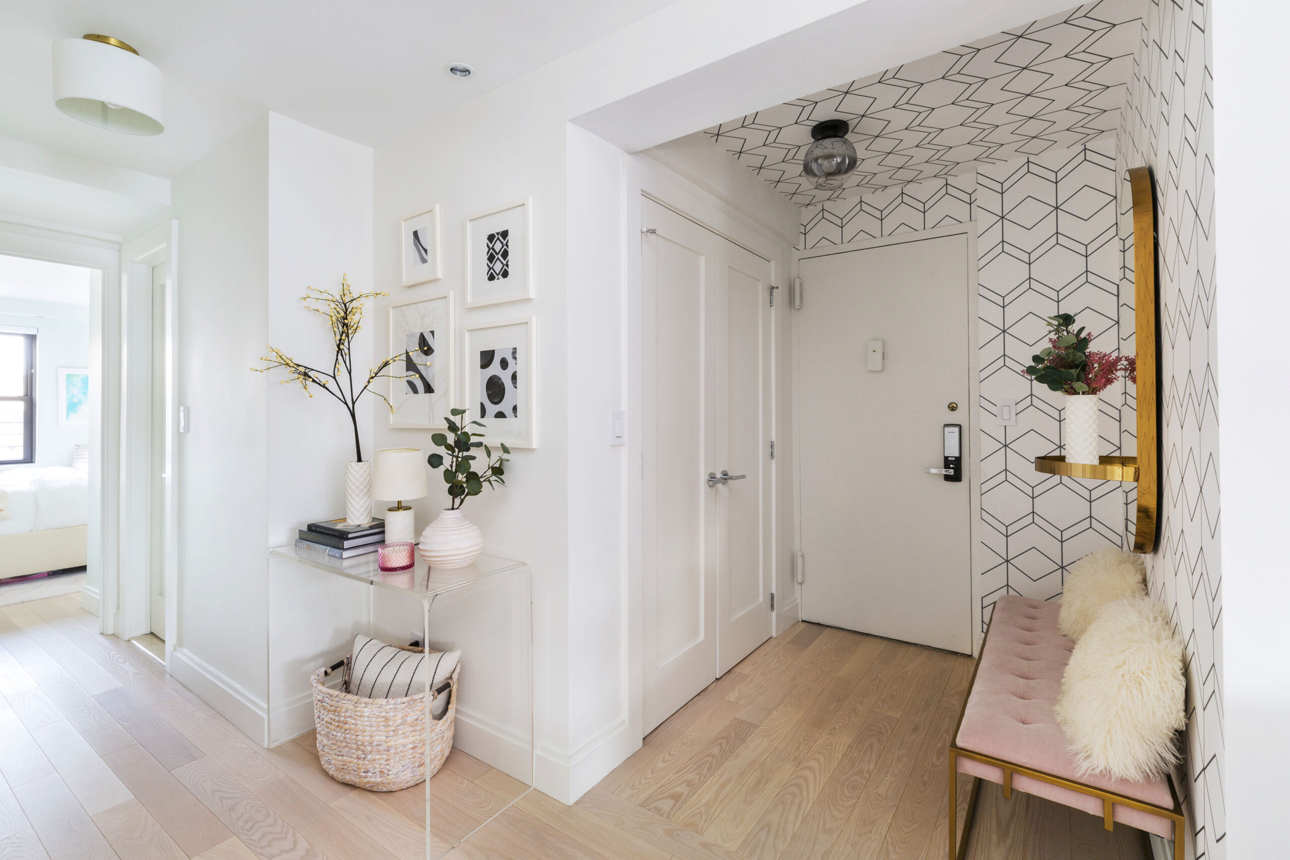 Upper East Side foyer with geometric wallpaper on the walls and ceiling, pink bench, brass shelf, round mirror, gallery art, and pale wood floors.