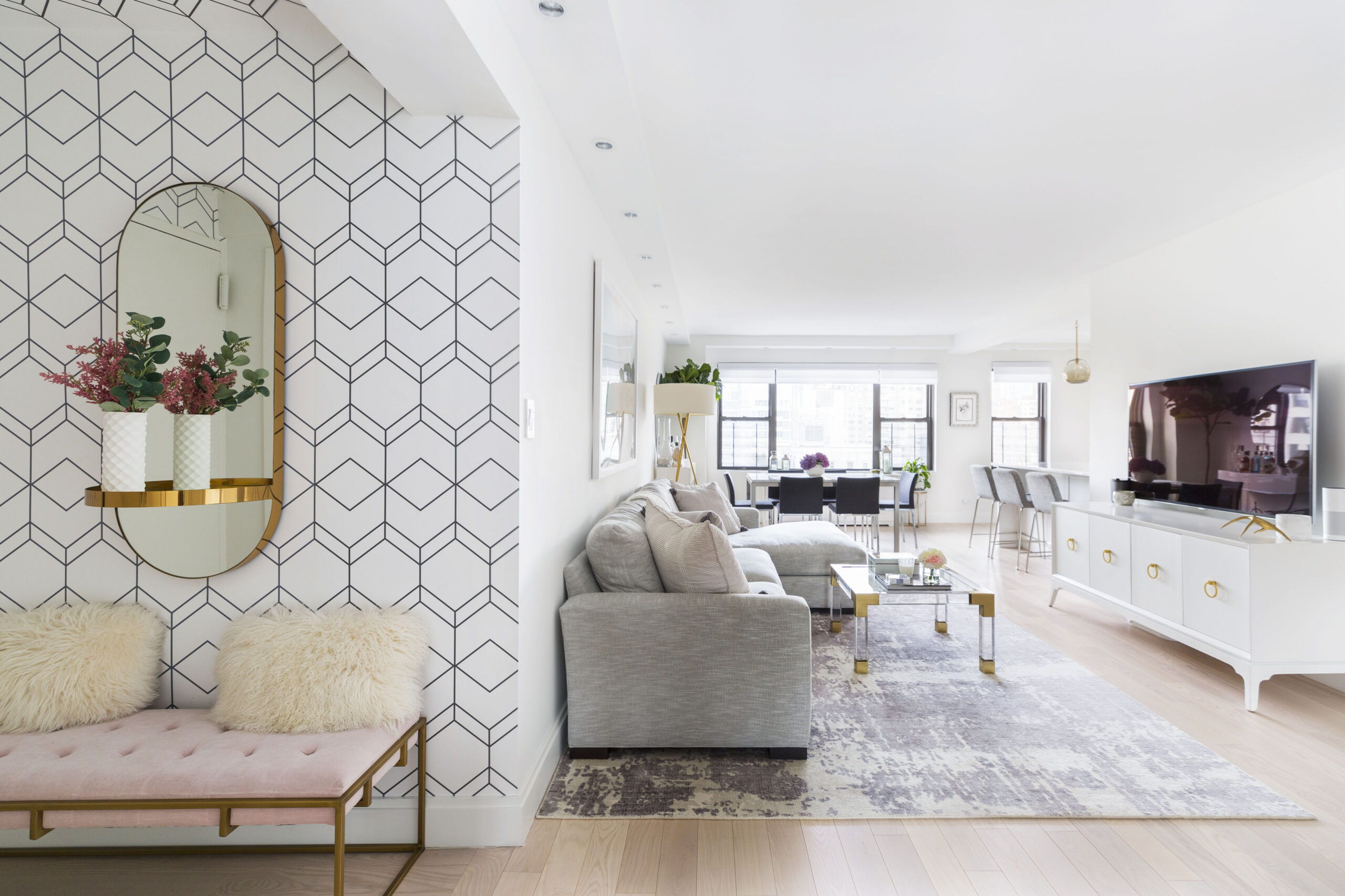Upper East Side foyer pairs geometric wallpaper, a pink bench, and brass mirror with open views to the living and dining area after the remodel.