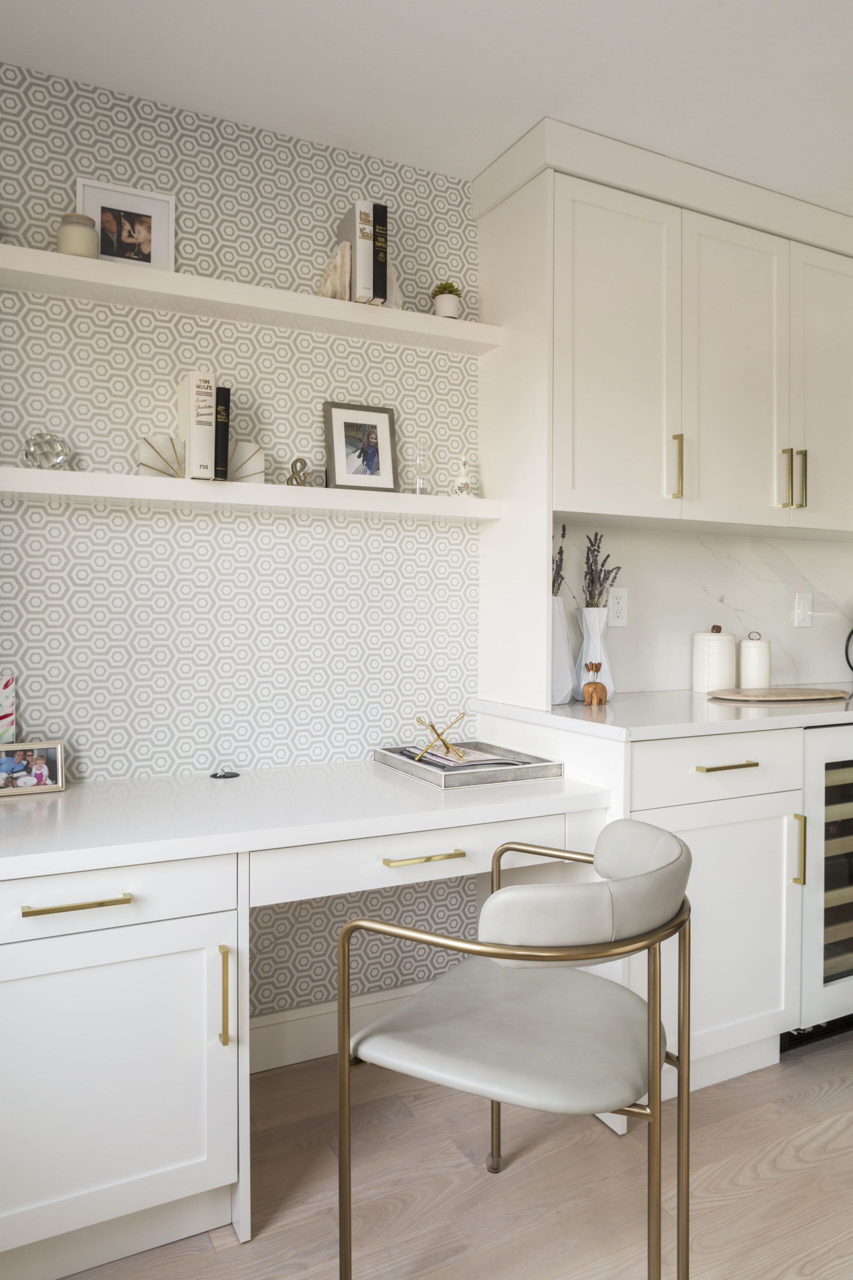 Upper East Side kitchen desk niche with patterned wallpaper sits beside white cabinets, brass pulls, island seating, and built-in kitchen storage.