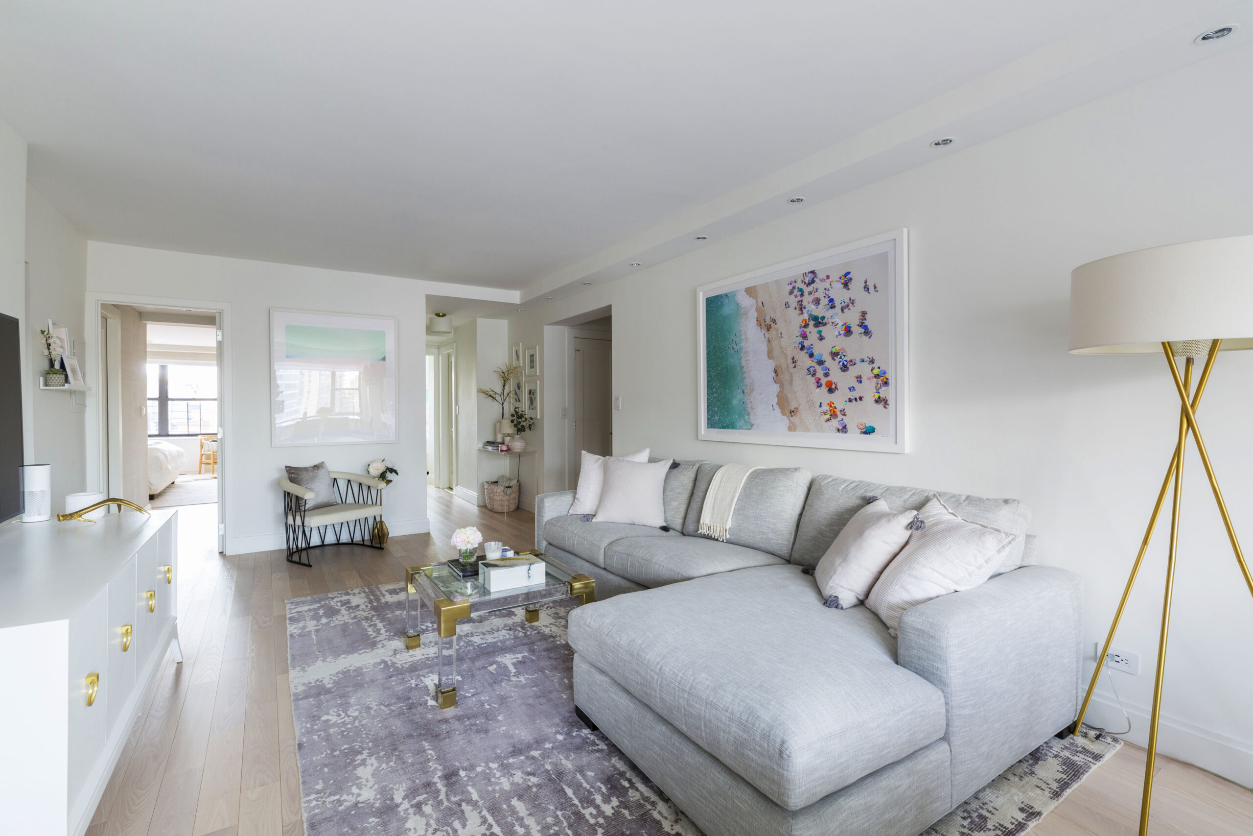 Upper East Side living area with pale wood floors, a gray sectional, brass floor lamp, beach artwork, and open hallway views after the co-op renovation.