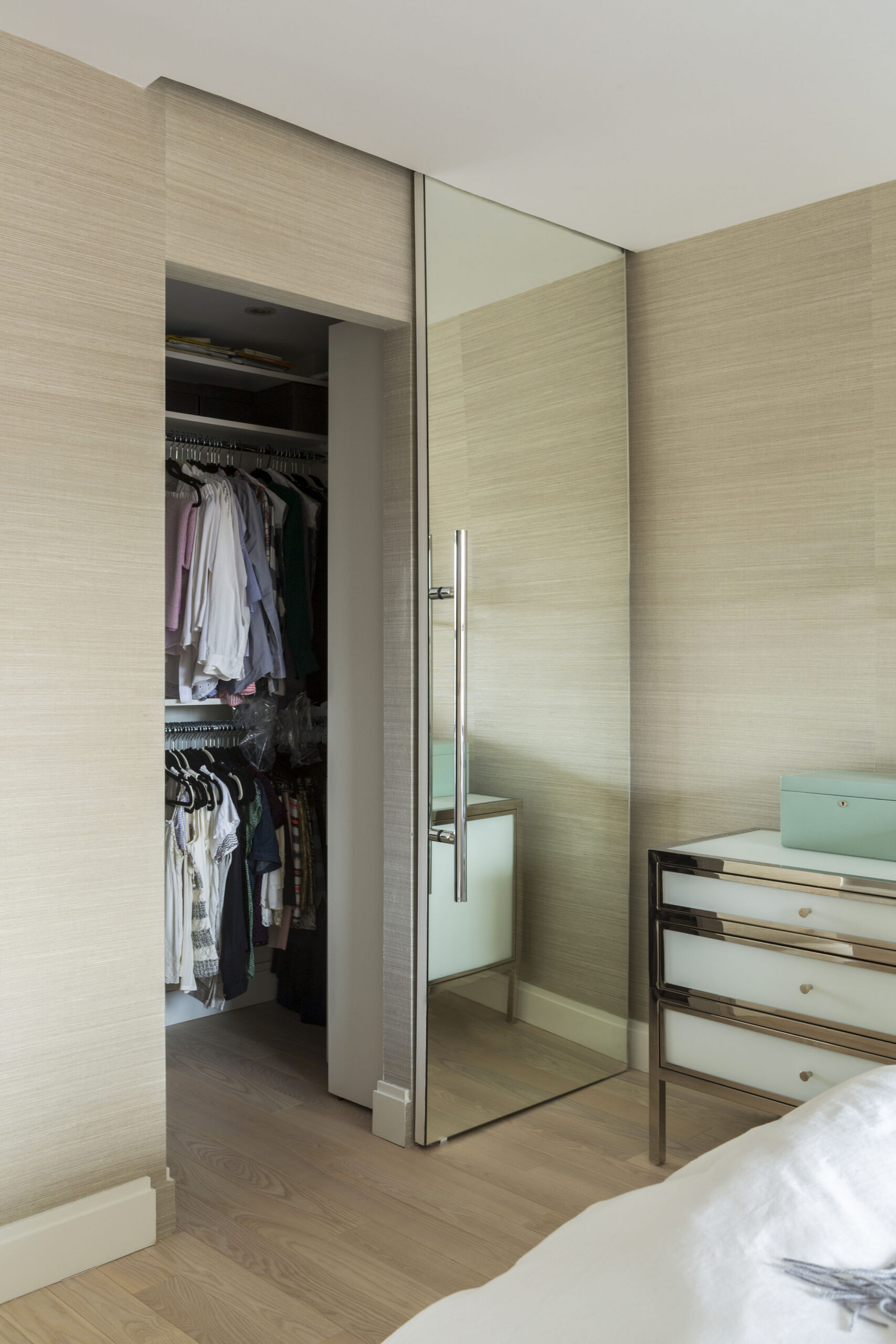 Upper East Side primary bedroom with grasscloth-style wallpaper, pale wood floors, a mirrored closet door, and added closet storage after the remodel.