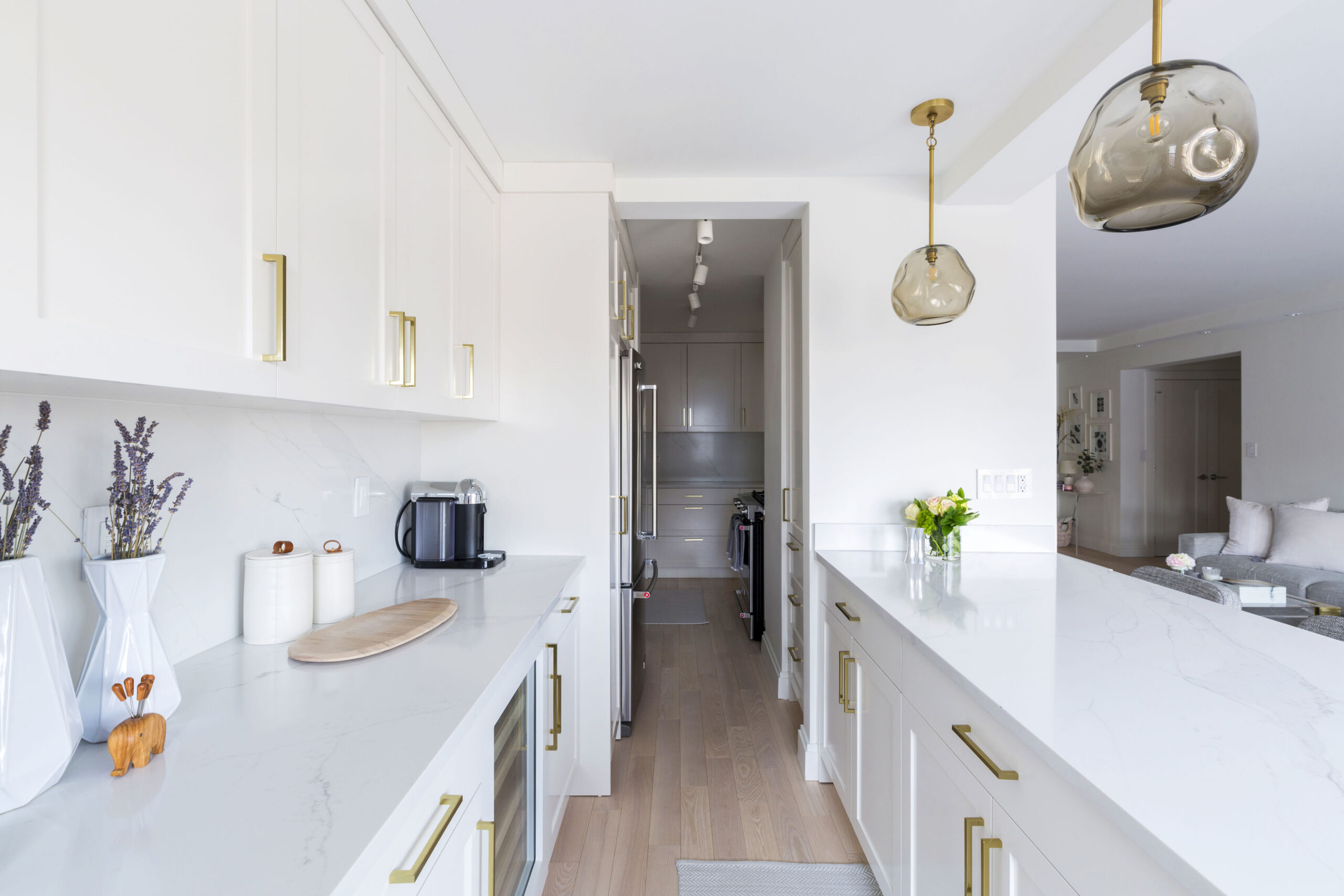 Renovated Upper East Side kitchen with white cabinets, brass pulls, marble-style counters, island storage, glass pendants, and an open living room view.