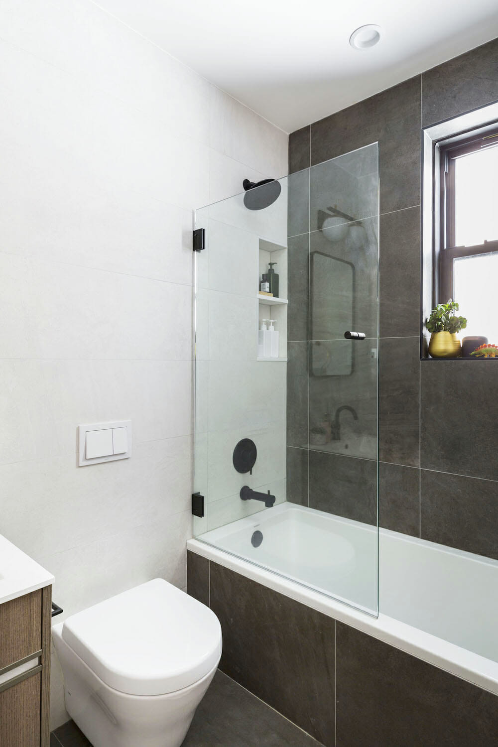 bathroom with dark gray tiles on floor and side of the bathtub and one shower wall and bathtub with black fixtures and recessed shelves and window after renovation