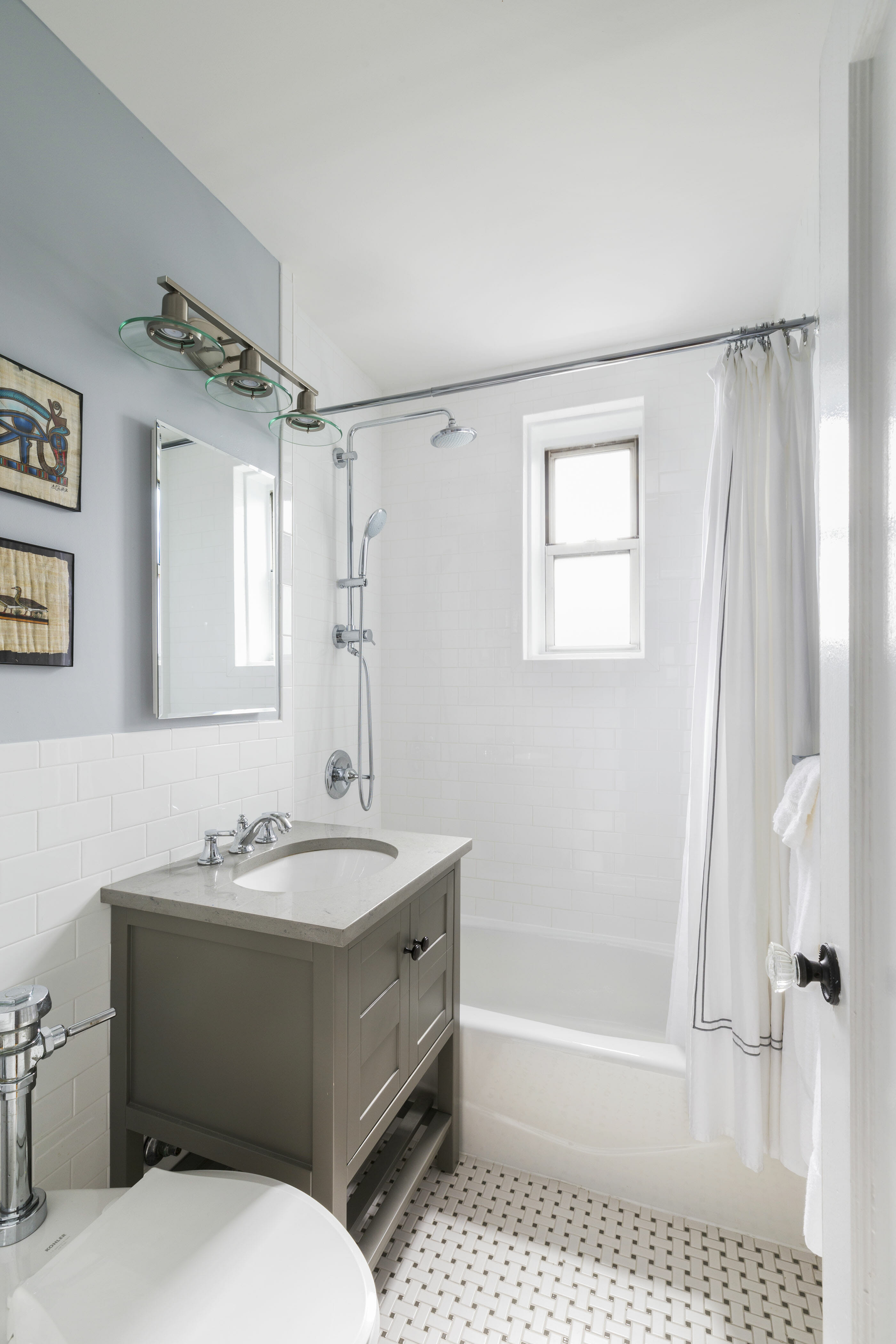 bathroom vanity with undermount porcelain sink and mirror and white tiles on half walls and gray walls and bathtub with a window and floor tiles after renovation
