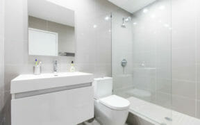 white floating bathroom vanity with farmhouse sink and mirror and toilet and walk-in shower with half glass wall and gray tiles on walls and floors after renovation