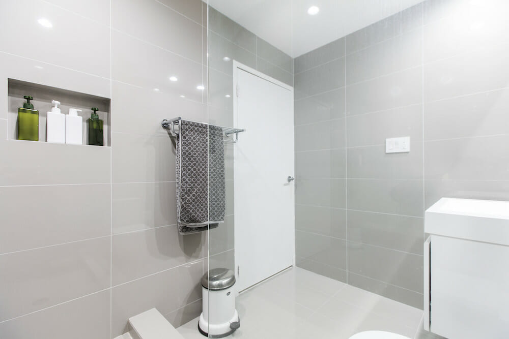 walk-in shower with half glass wall and recessed shelf on wall and light gray tiles on walls and floors  and white vanity after renovation