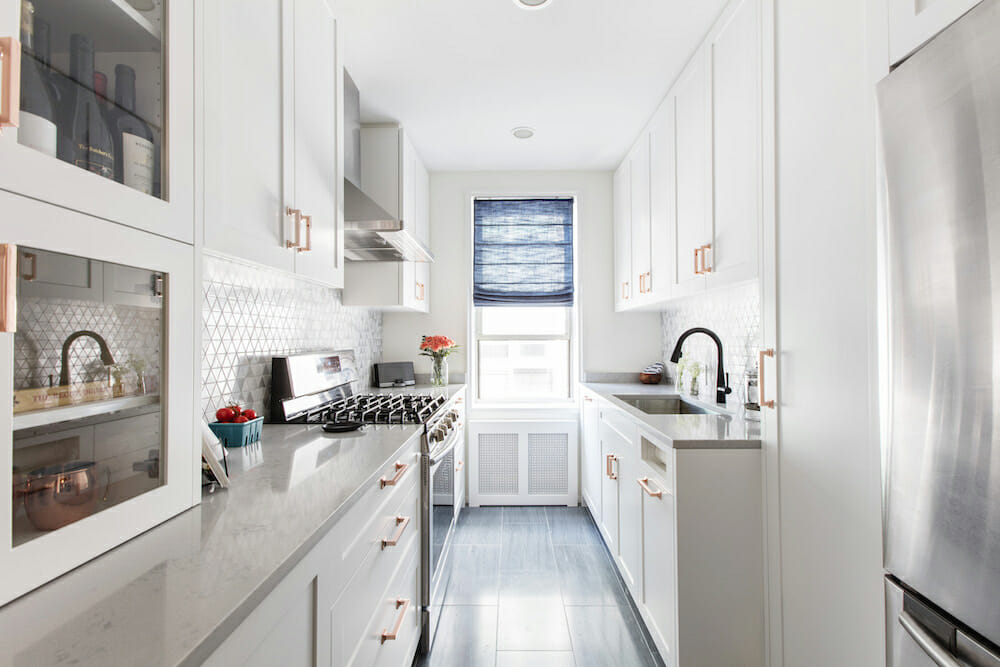 white kitchen cabinets with copper handles and overhead cabinet with glass doors and marble countertop and gas cooking range with hood after renovation
