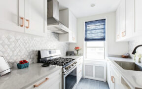 white kitchen cabinets with copper handles and gas cooking range with hood and dark gray floor tiles and radiator with cover below window after renovation