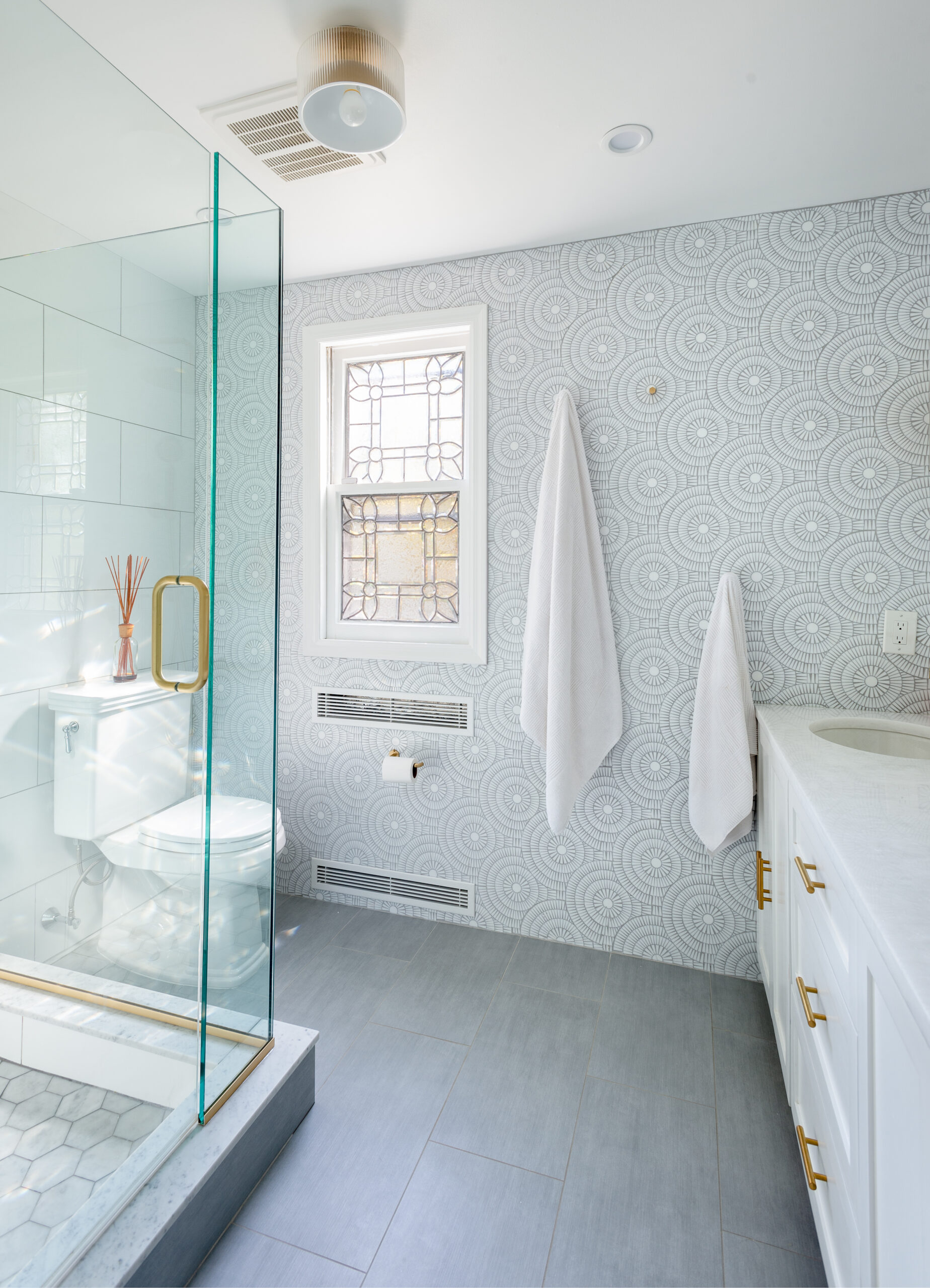 Westchester bathroom with a glass shower, patterned wall covering, gray floor tile, brass accents, a white vanity, and an original stained-glass window.
