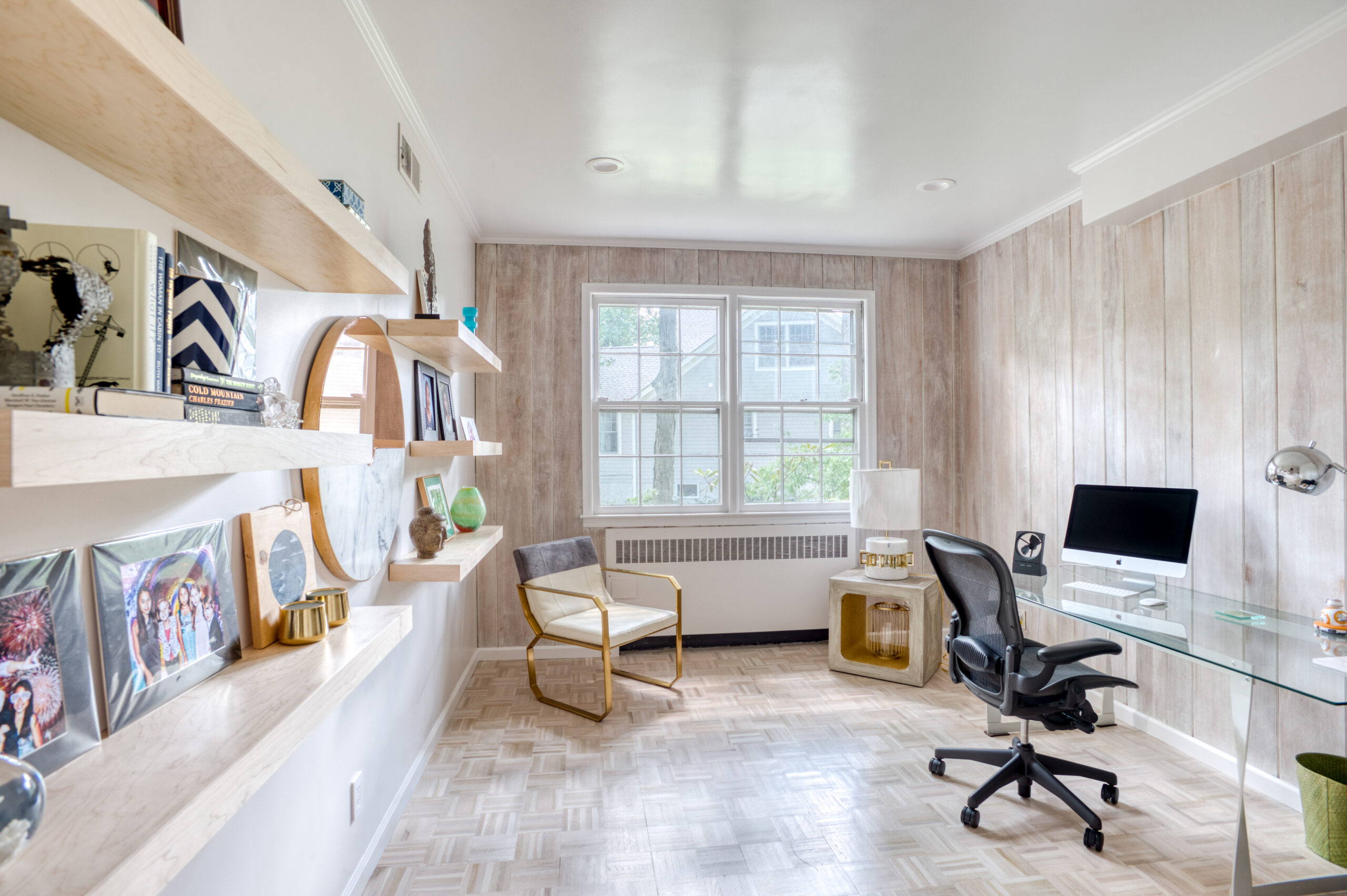 Westchester home office in a modern traditional home renovation with restored white-stained parquet, floating shelves, a glass desk, and wood-look walls.