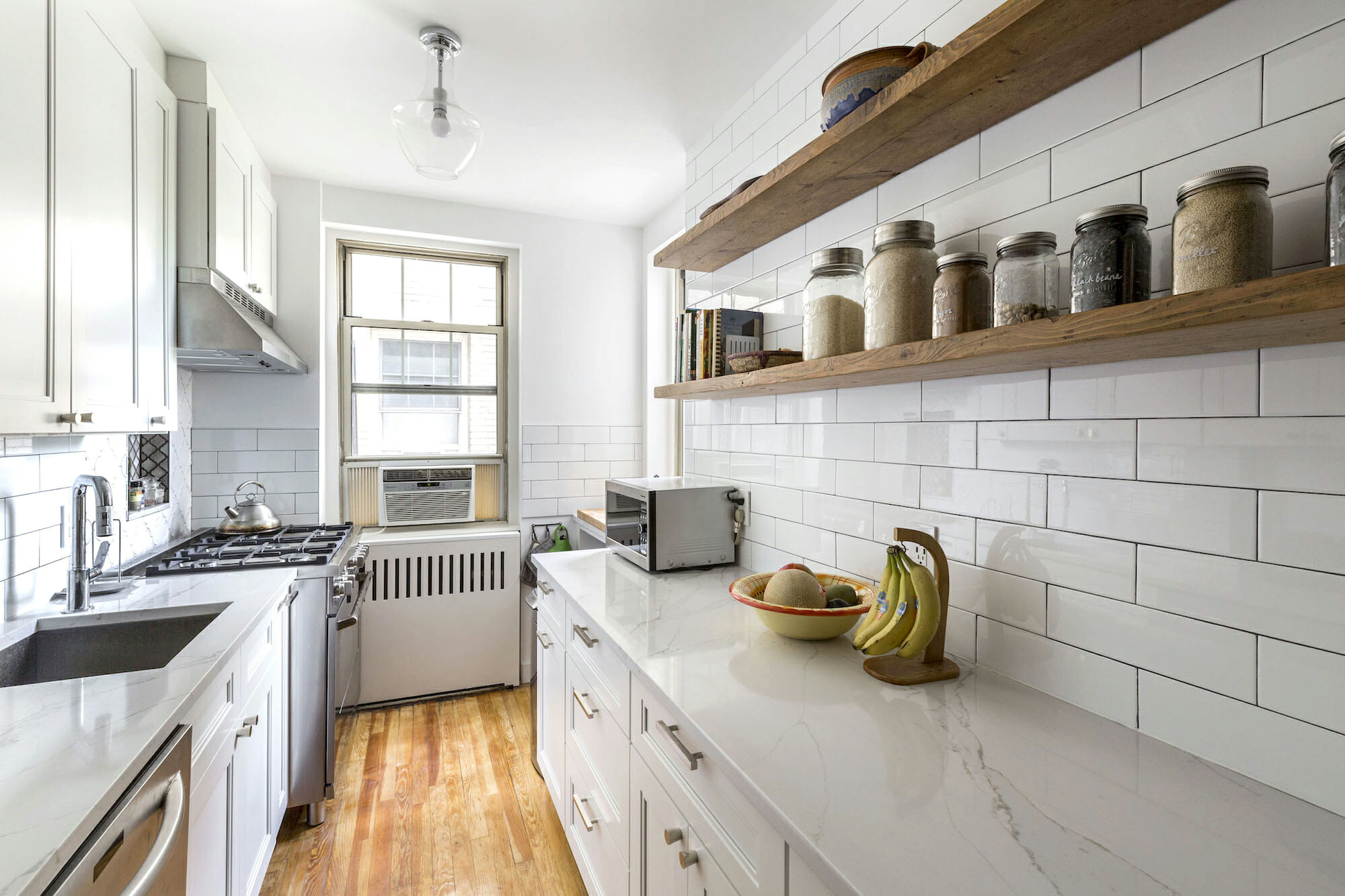 white kitchen cabinets with subway tiles on walls and marble countertop with farmhouse sink and stainless steel appliances and floating wooden shelves after renovation