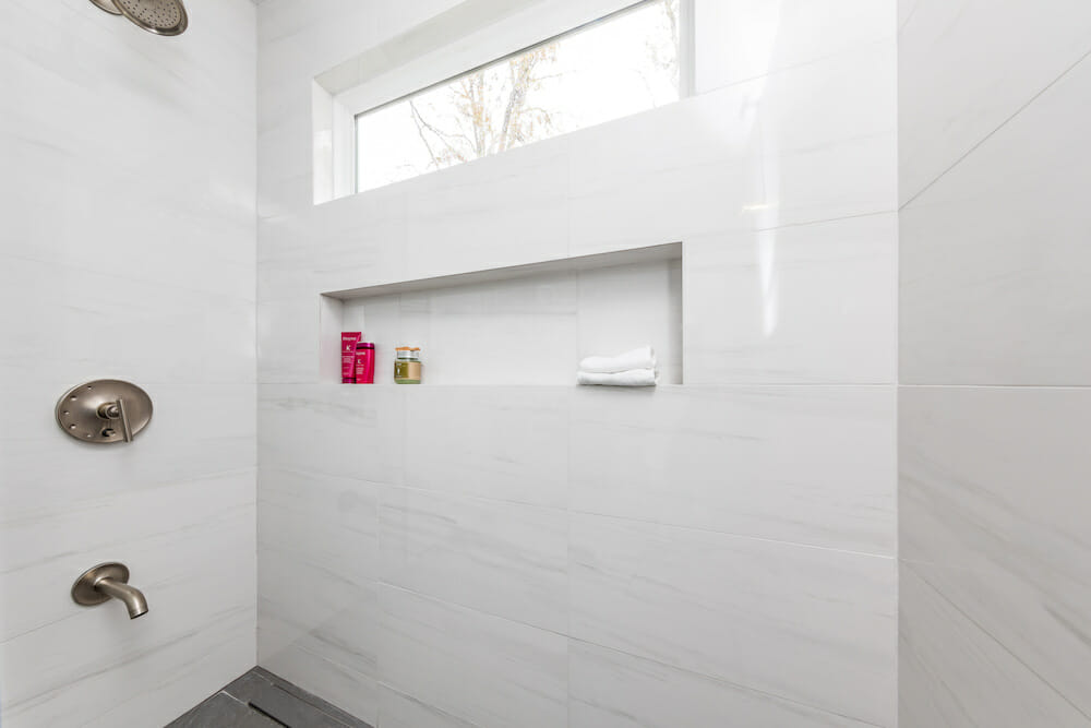 walk in shower with marble tile walls and built in shelf and a small overhead window after renovation