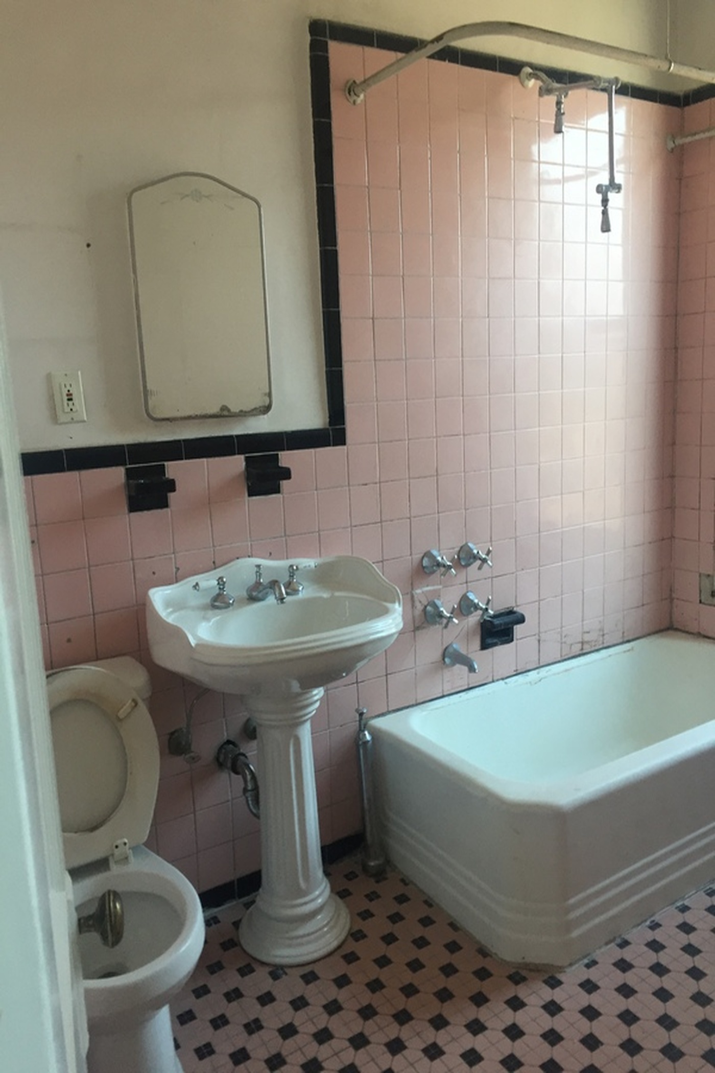 Boerum Hill bathroom before the remodel with pink wall tile, black trim, a pedestal sink, built-in tub, exposed fixtures, and mosaic floor tile.