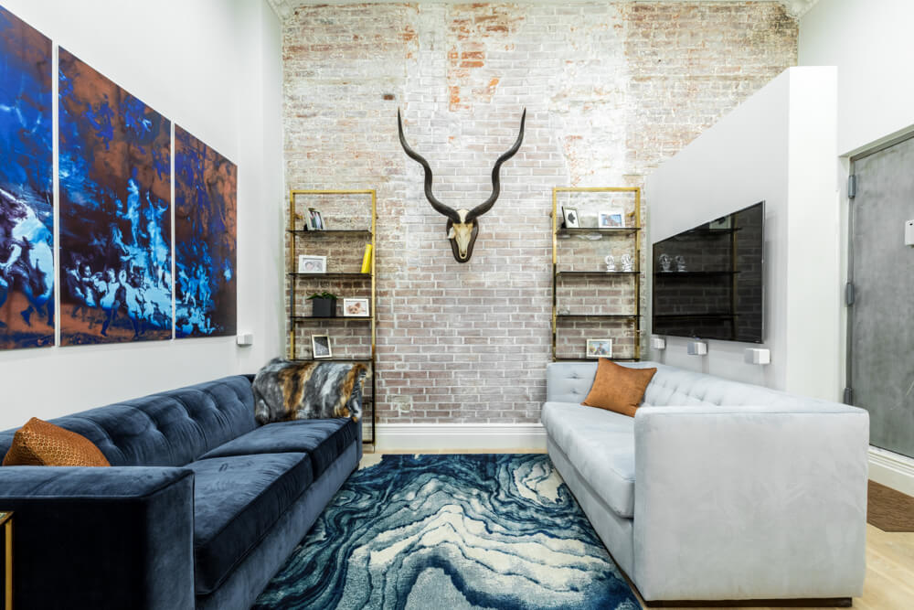 light brick wall in a narrow living room with plush gray and blue furniture and open metal shelves after renovation