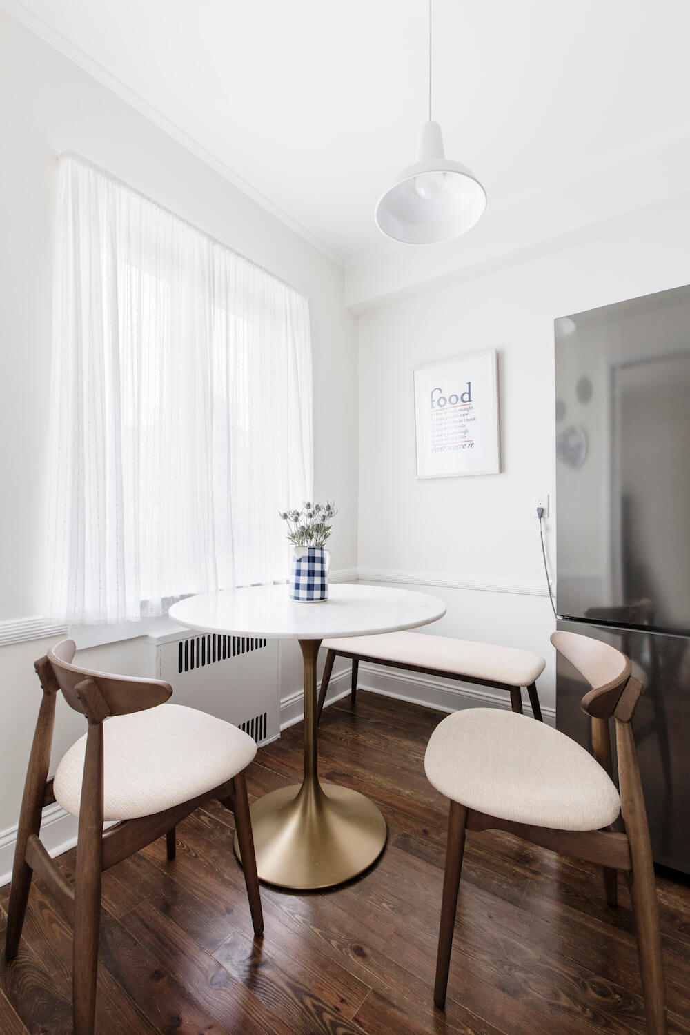 breakfast nook with round white table and chairs and a bench on wooden floor and white pendant light on top and large window with white sheer curtain after renovation
