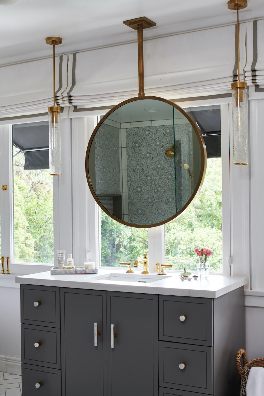 large gray single vanity with white marble countertop and brushed nickel hardware and gold faucet and fixtures and hanging circular mirror infront of window after renovation