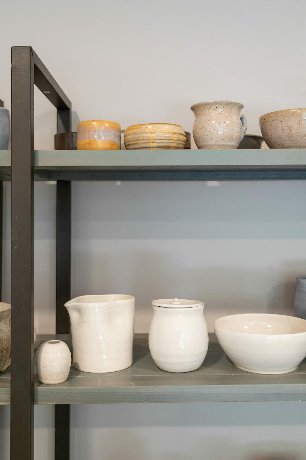 Grey and black minimal storage unit displaying handcrafted white and brown ceramic cups, saucers and beakers