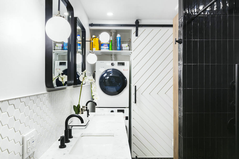 double vanity sinks with marble countertop and black faucet and fixtures and mirrors with black frames and shower walls with black subway tiles and stacked washer dryer with a chevron design white barn door after renovation