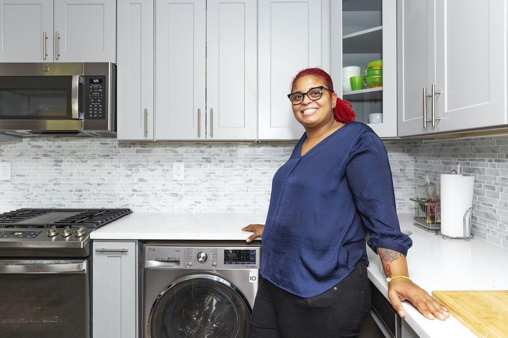 sally posing in front of washing machine in her kitchen with white cabinets and white countertop and stainless steel appliances after renovation