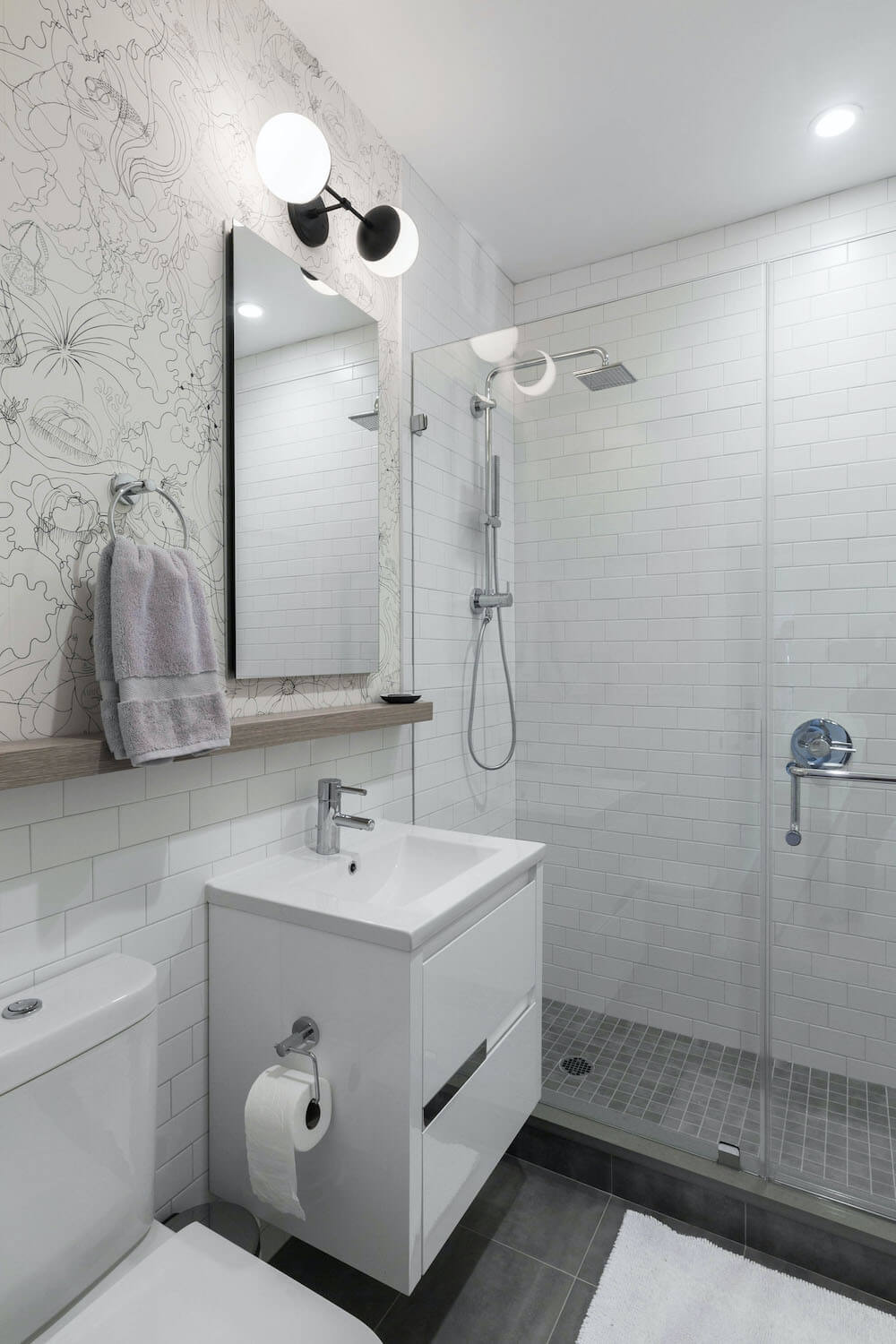 bathroom vanity with mirror and wall mounted light and wood trim and walk-in shower with white subway wall tiles and glass door and chrome shower head and fixtures and dark gray floor tiles after renovation