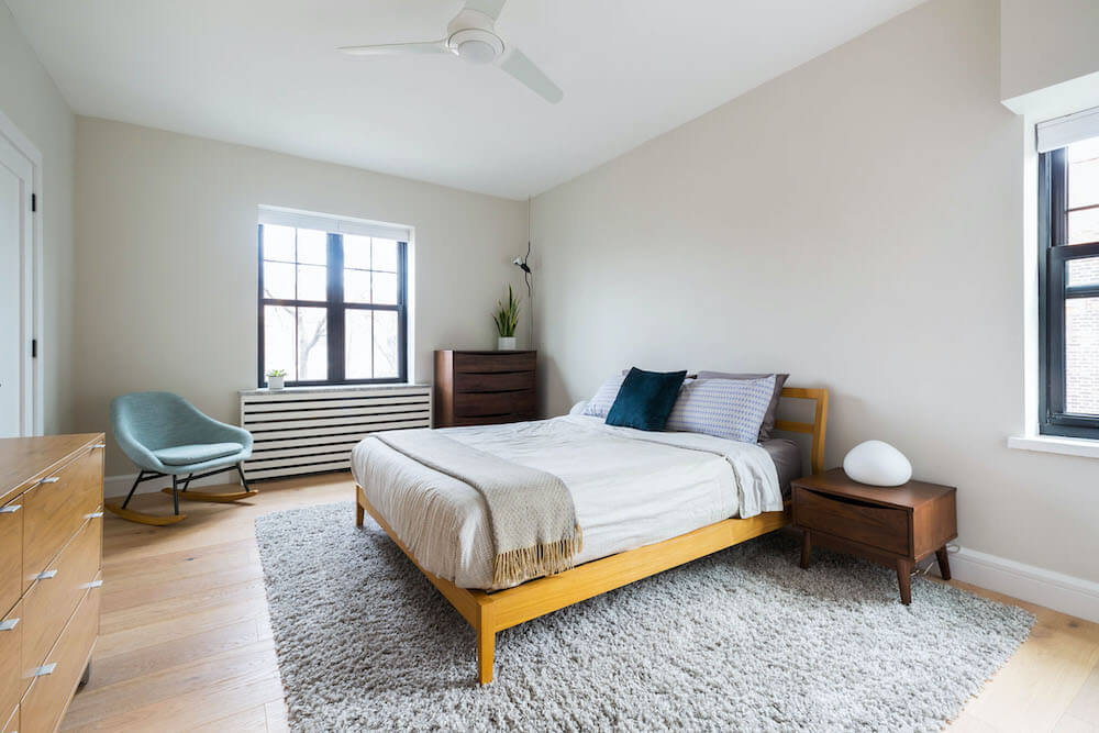 bedroom with beige walls and white trims and hardwood floors and radiator with cover under window with black frame and ceiling fan after renovation