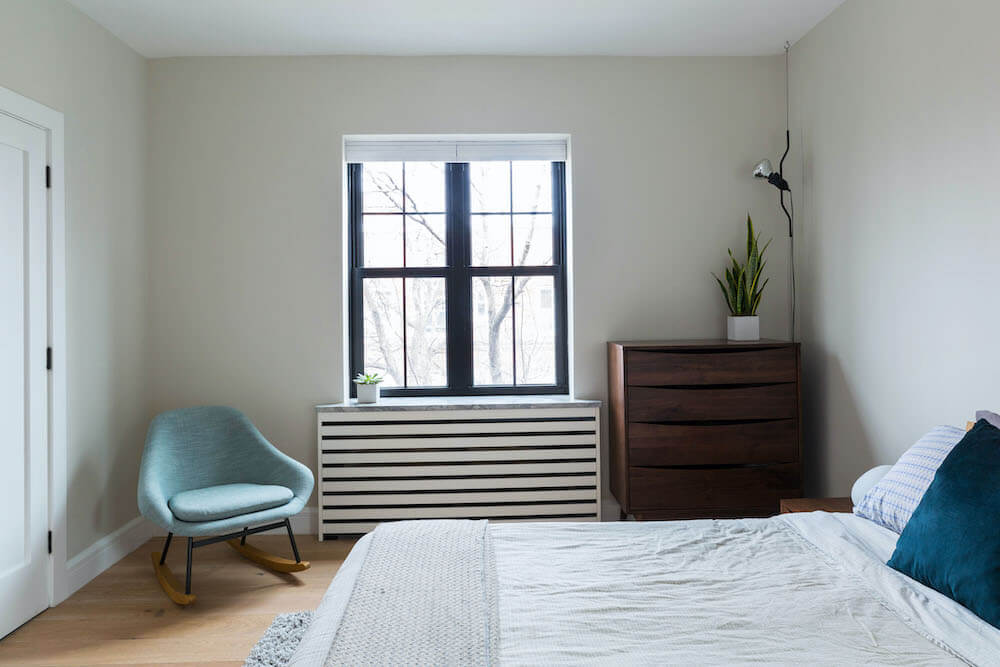 bedroom with beige walls and white trims and hardwood floors and radiator with cover under window with black frame after renovation