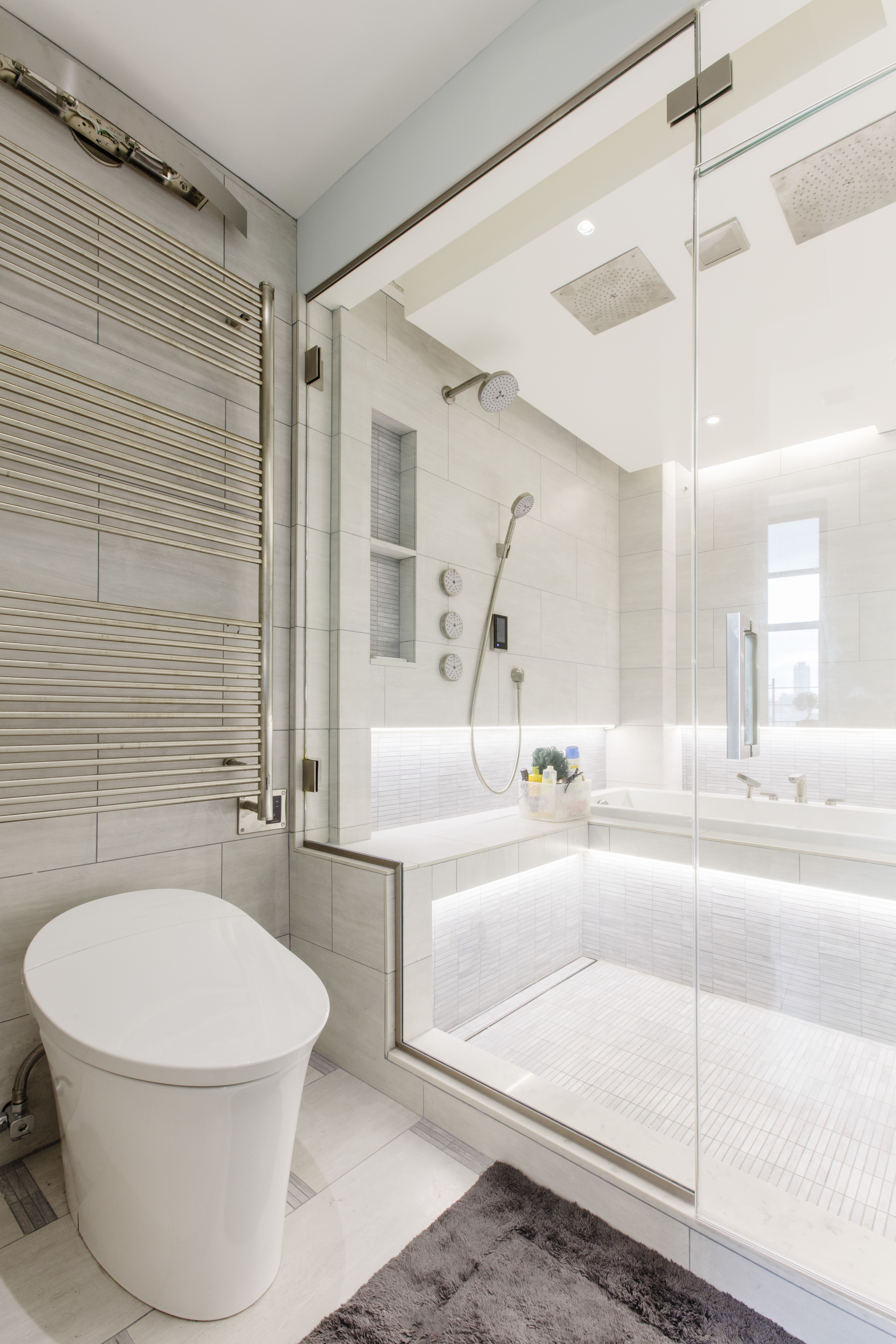 Glass-enclosed shower bench in Central Park West with dual rain heads, a heated towel rack, inset tub, pale tile, and illuminated wraparound seating.