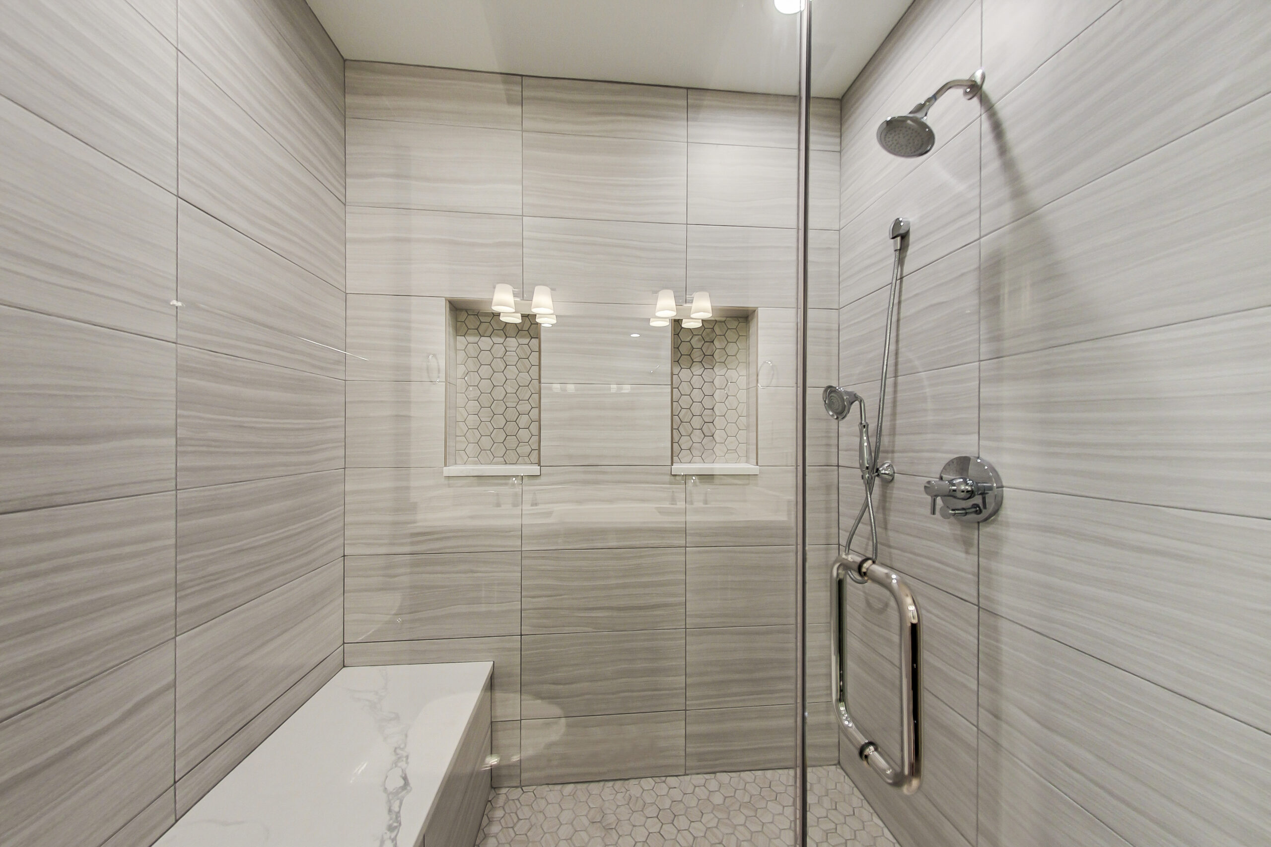 Long shower bench in a Chicago walk-in shower with large-format gray tile, twin hexagon niches, glass panels, and handheld shower fixtures.