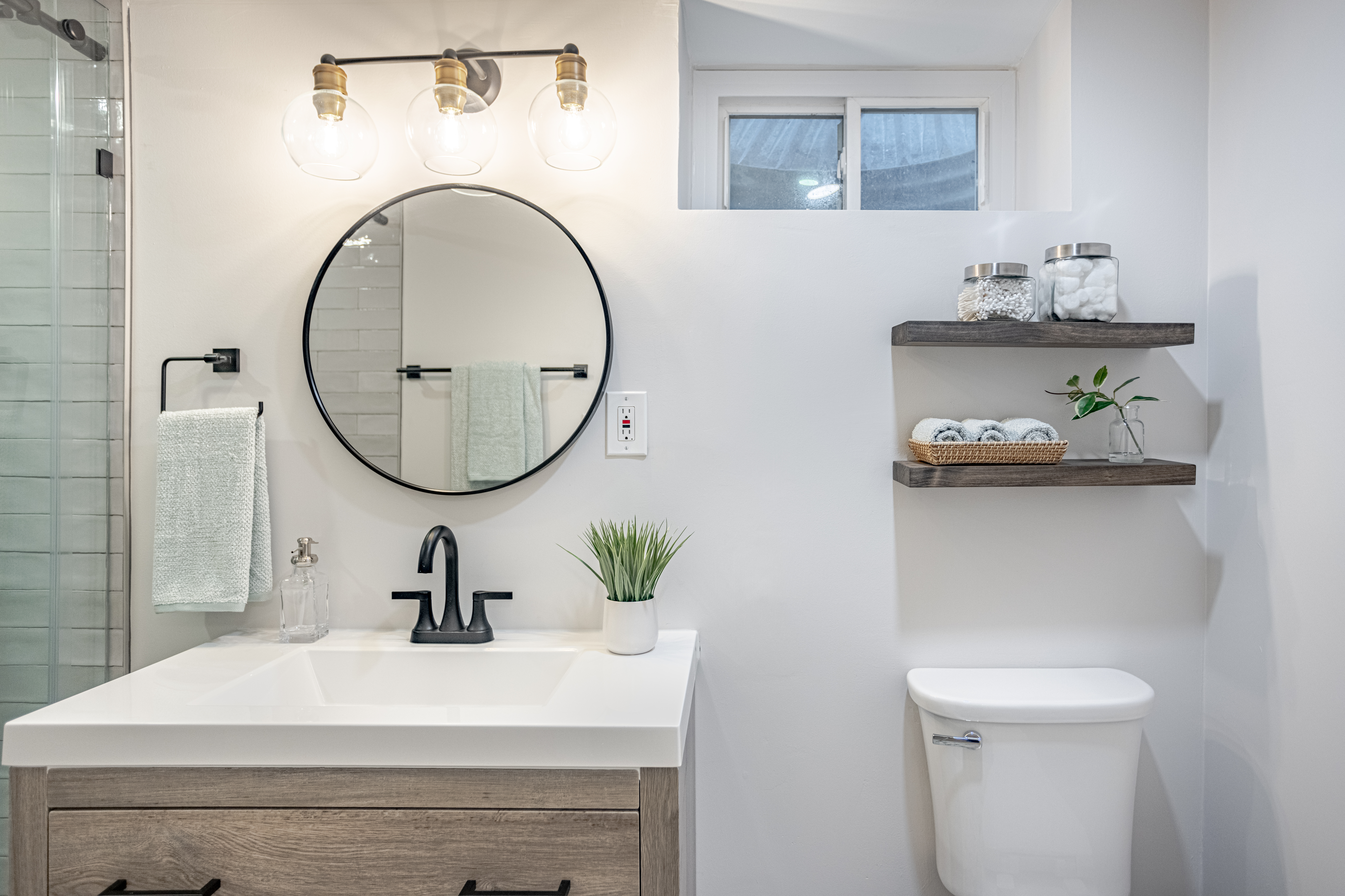 Arlington basement bathroom remodel pairs a wood vanity, round black mirror, floating shelves, bright lighting, and a small window for added daylight.