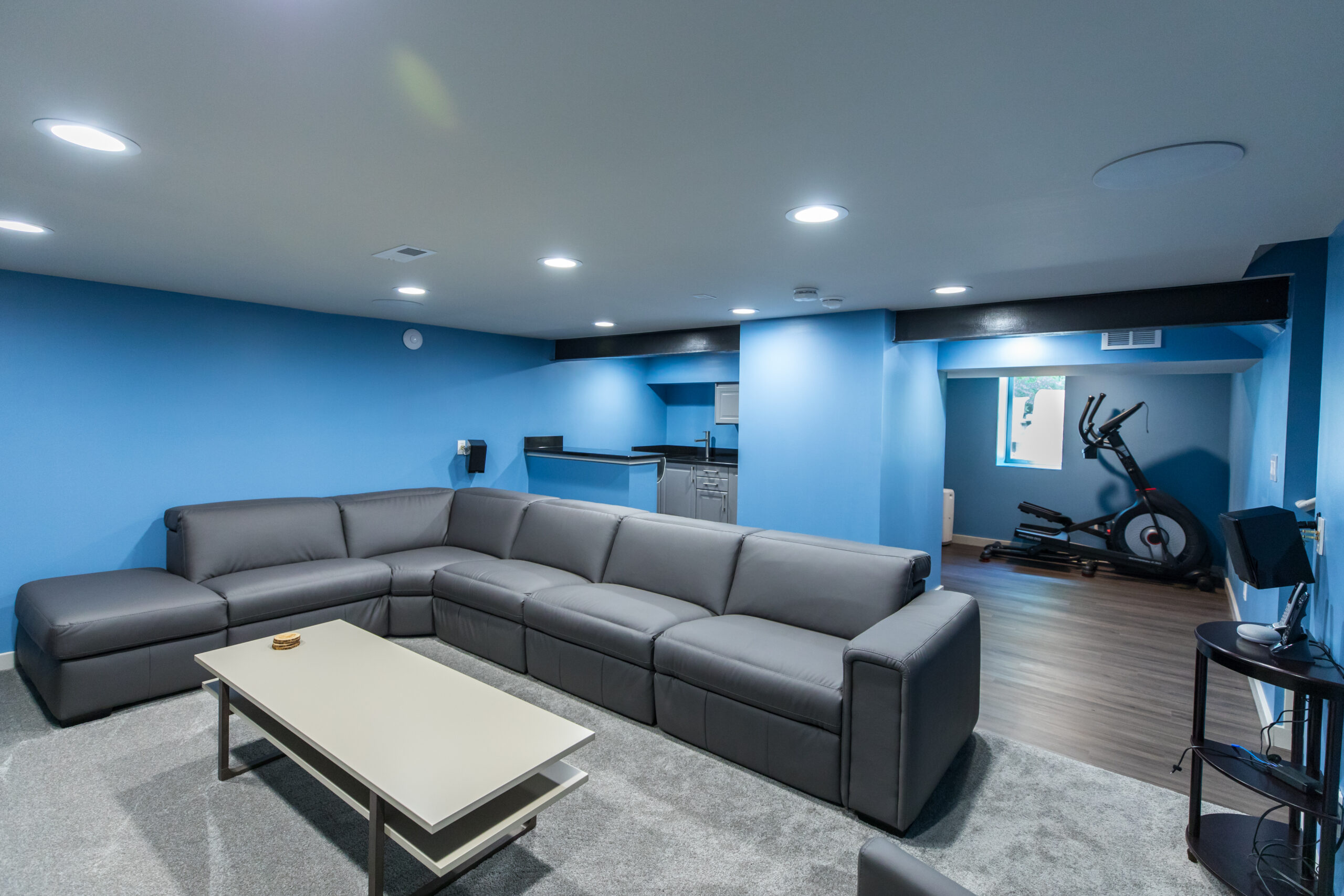 Philadelphia basement remodel combines a carpeted lounge, gray sectional, recessed lighting, bar area, vinyl plank flooring, and a dedicated fitness zone.