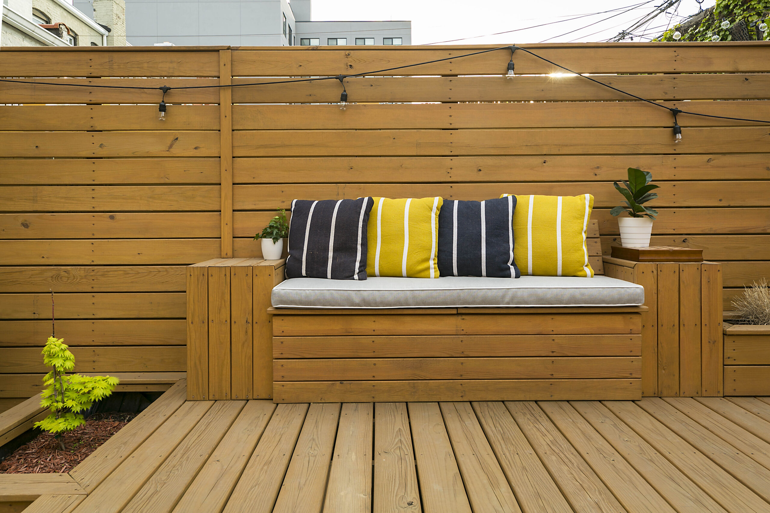 Brooklyn outdoor lighting design adds string lights above a built-in wood bench, privacy fencing, deck boards, and integrated planter boxes.
