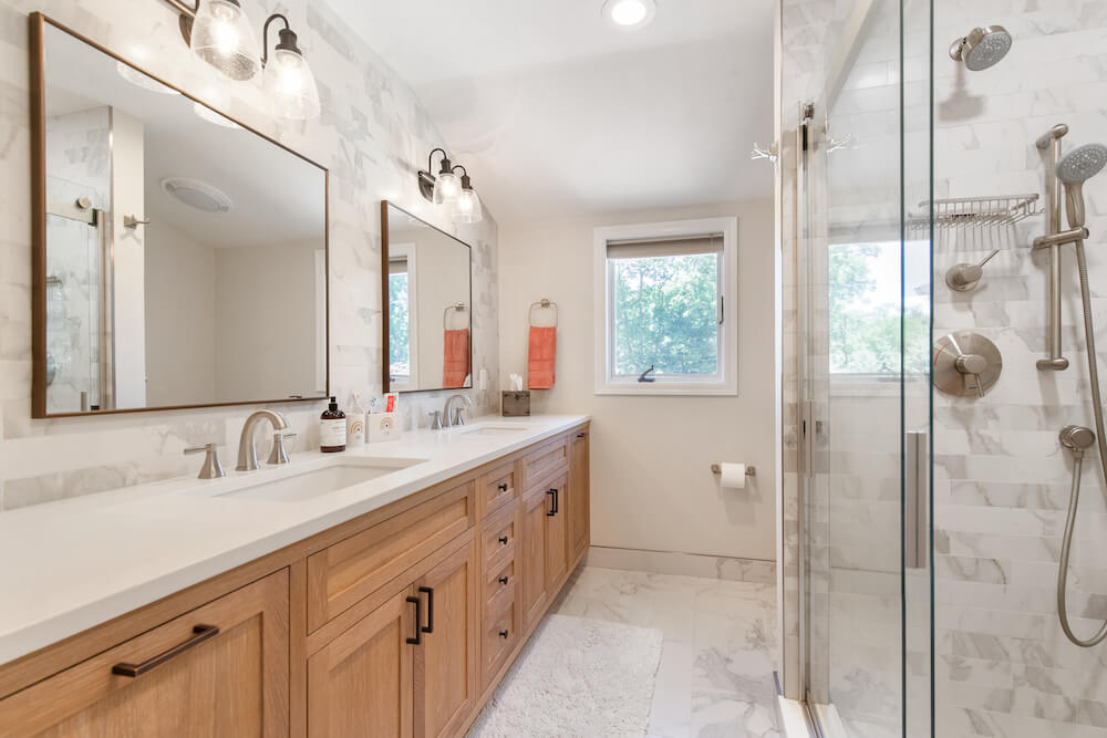 Harrison home lighting design pairs lit vanity fixtures and recessed lights with a double wood vanity, glass shower, and marble-look tile.