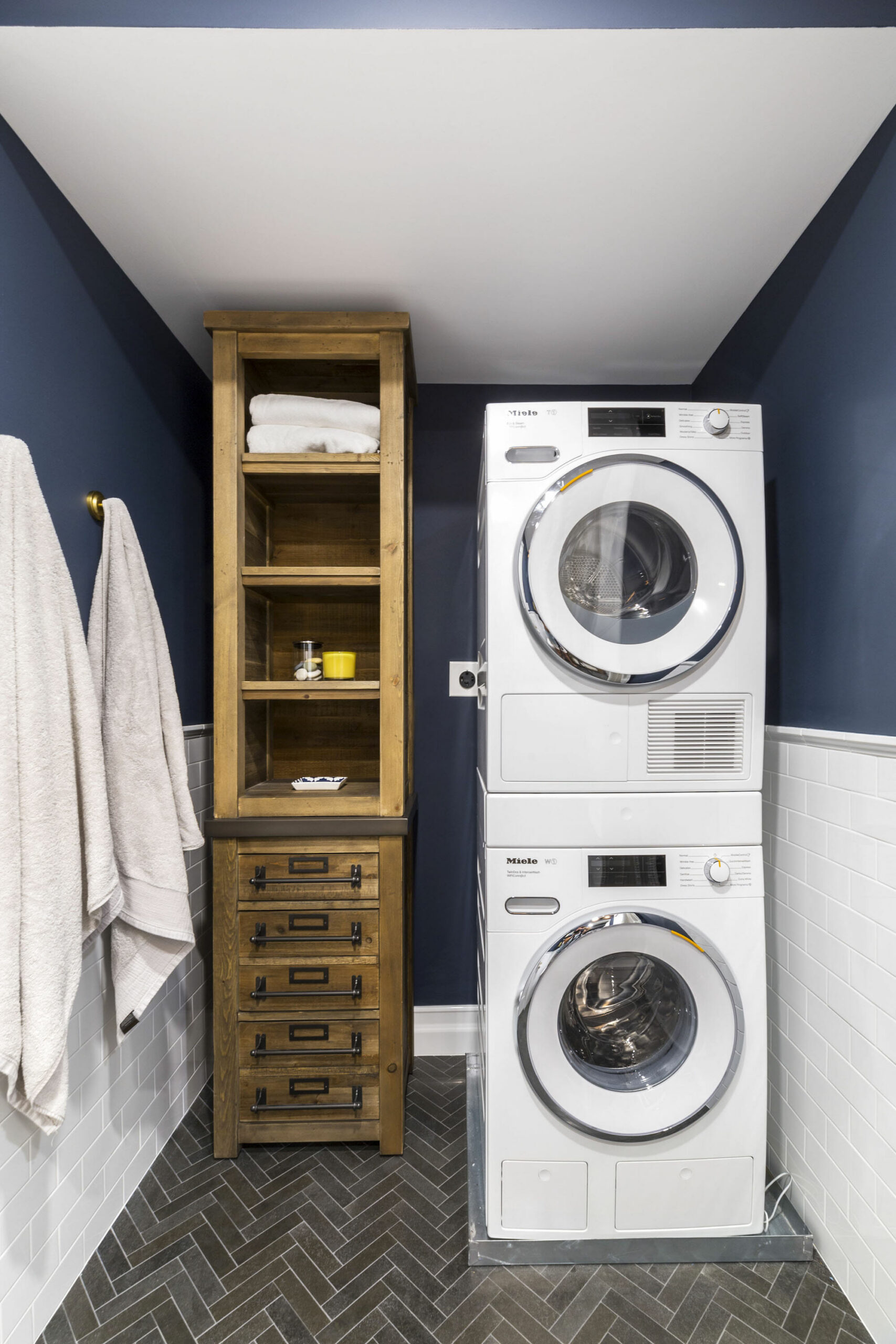 Washer and dryer installation in DUMBO stacks front-load units beside slim wood storage, navy walls, white subway tile, and herringbone flooring.