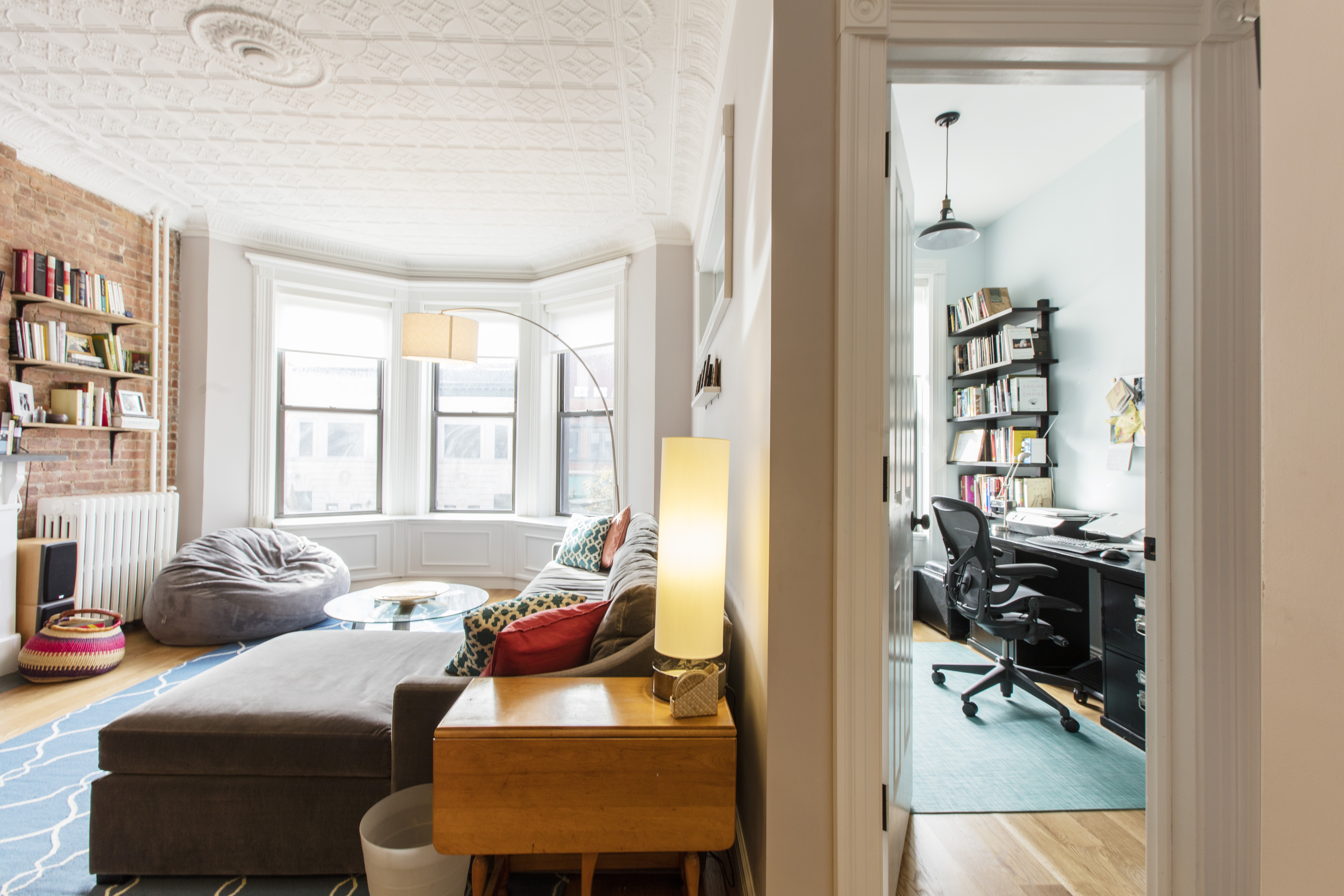 Brooklyn brownstone living room with exposed brick, tin ceiling, bay windows, oak floors, and a separate home office visible through the doorway.
