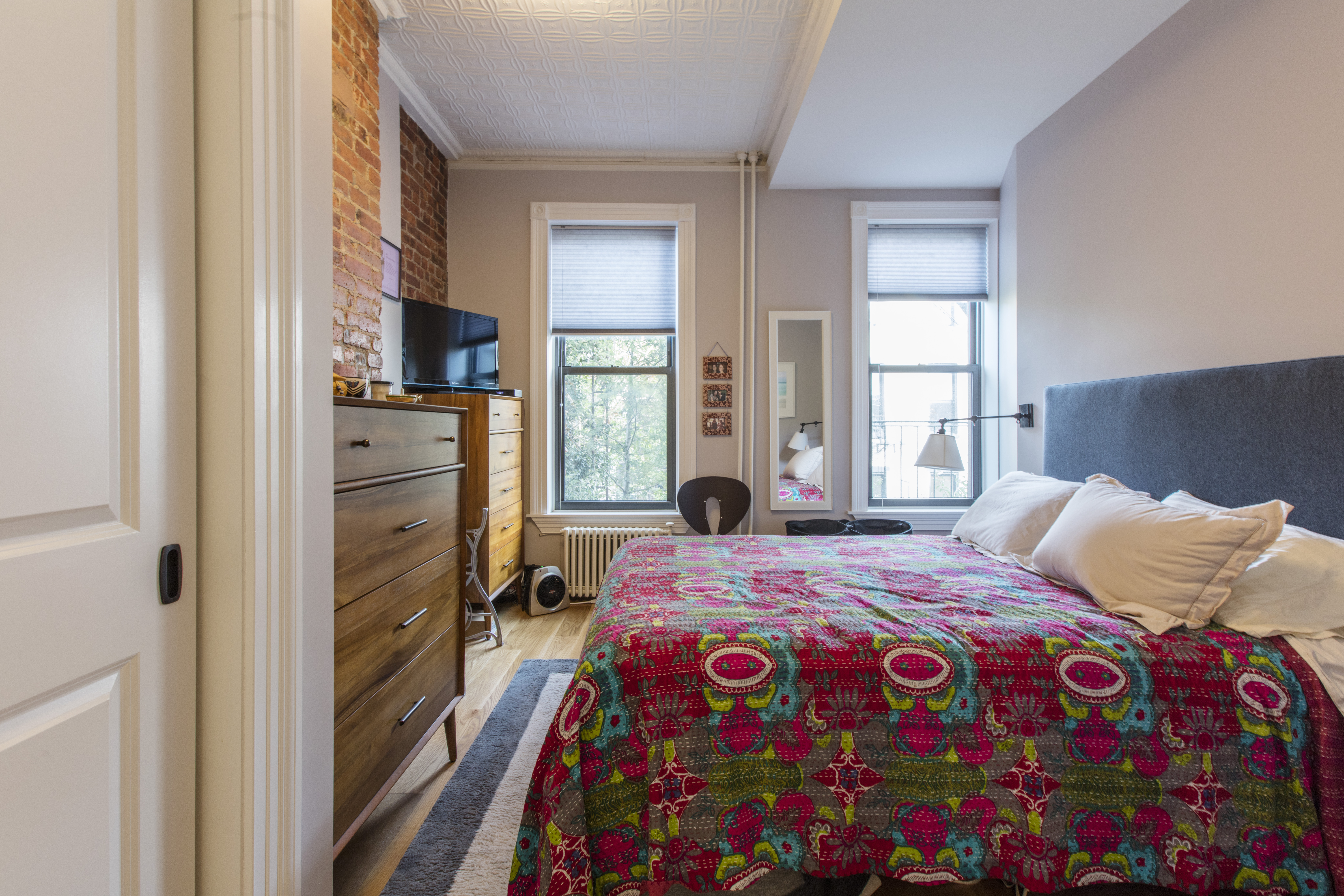Park Slope brownstone remodel bedroom with exposed brick, preserved tin ceiling, oak flooring, tall windows, and a simple modern bed and wood dressers.