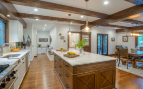 Renovated Tudor-style kitchen with white cabinets, oak island and beams, marble counters, and view to mudroom bench and dining table.