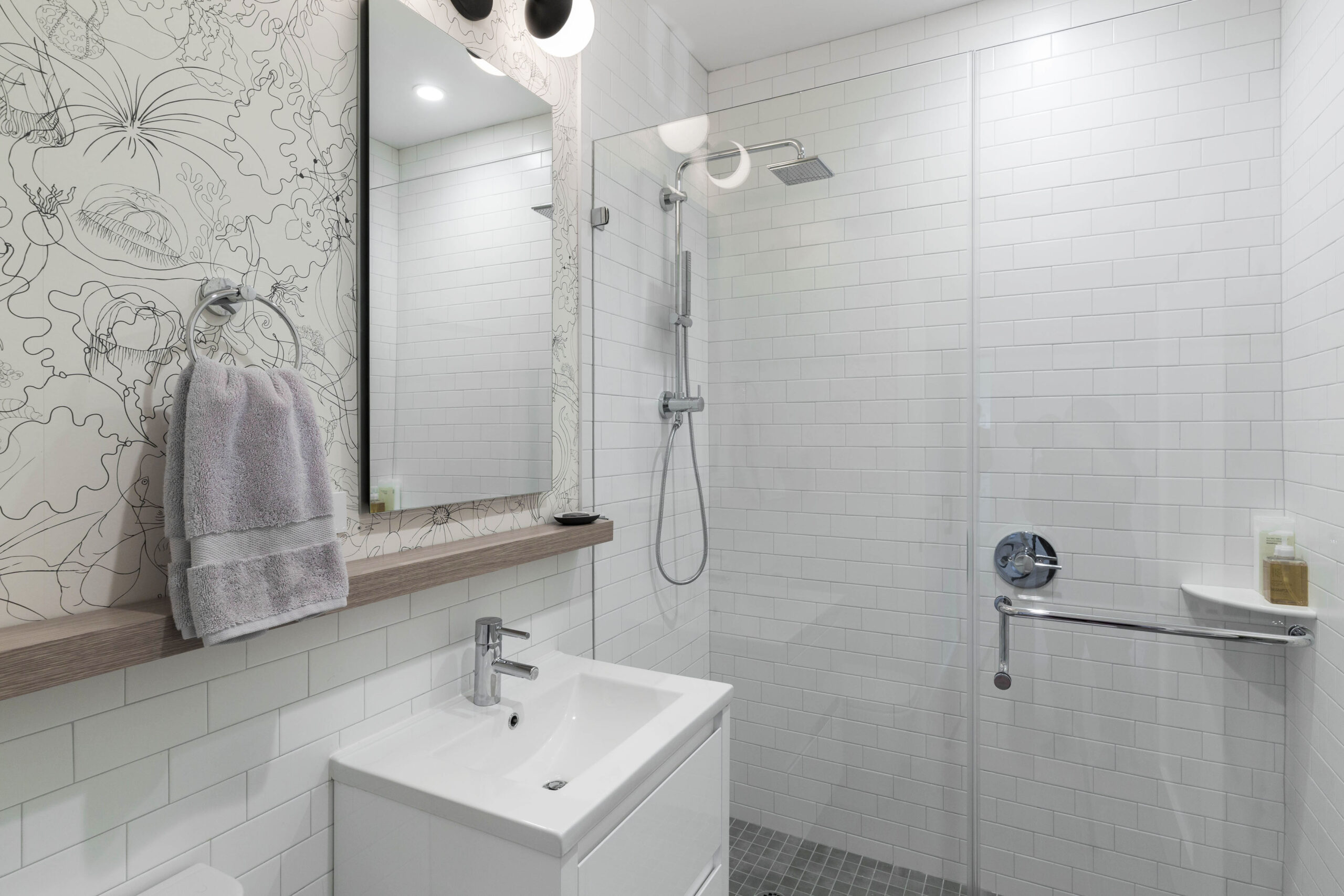 Walk-in shower with white subway tile, frameless glass, grab bar, rain showerhead, and a modern vanity with patterned wall detail.