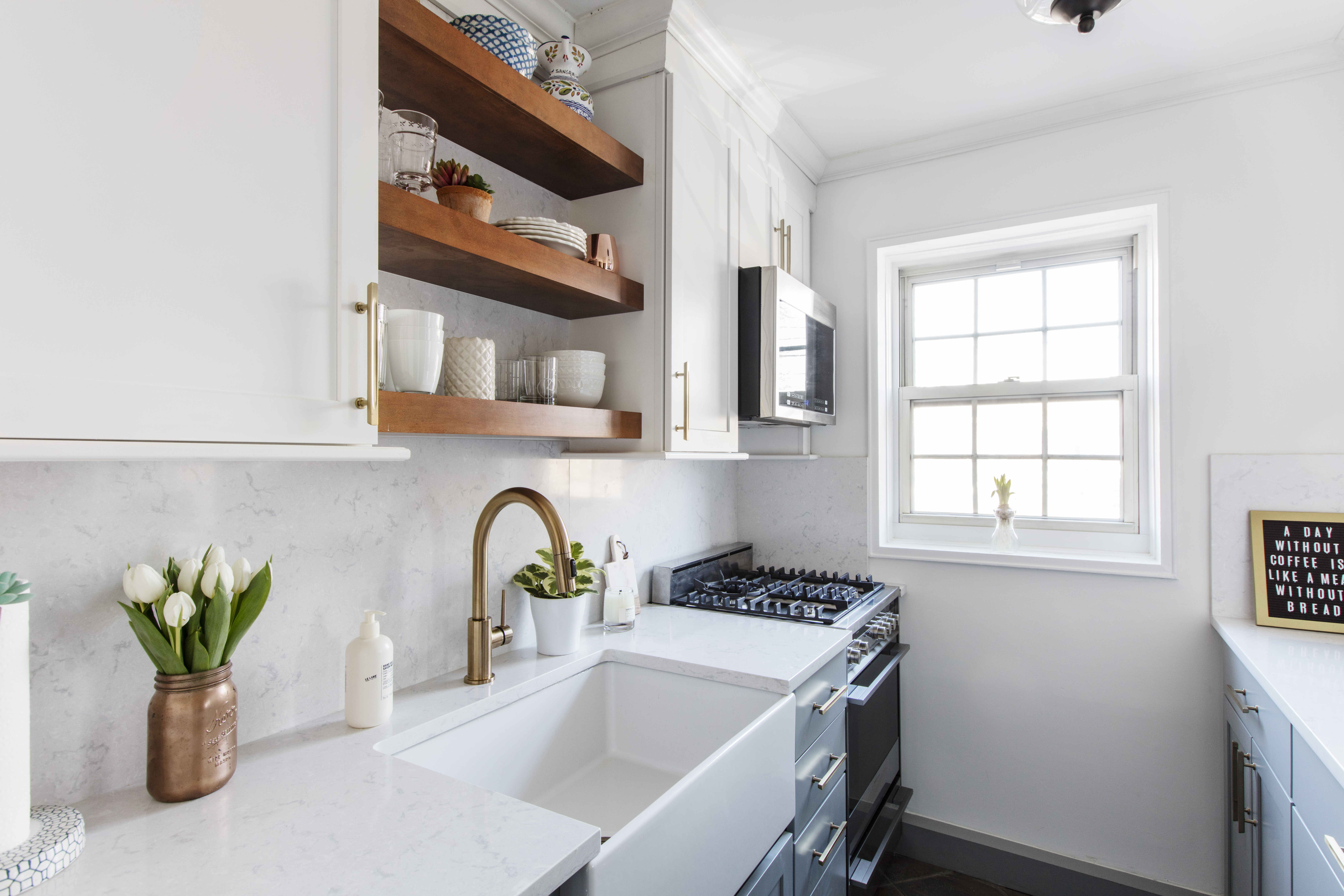 Galley kitchen with white uppers, brass faucet, open wood shelves, farmhouse sink, and a window.