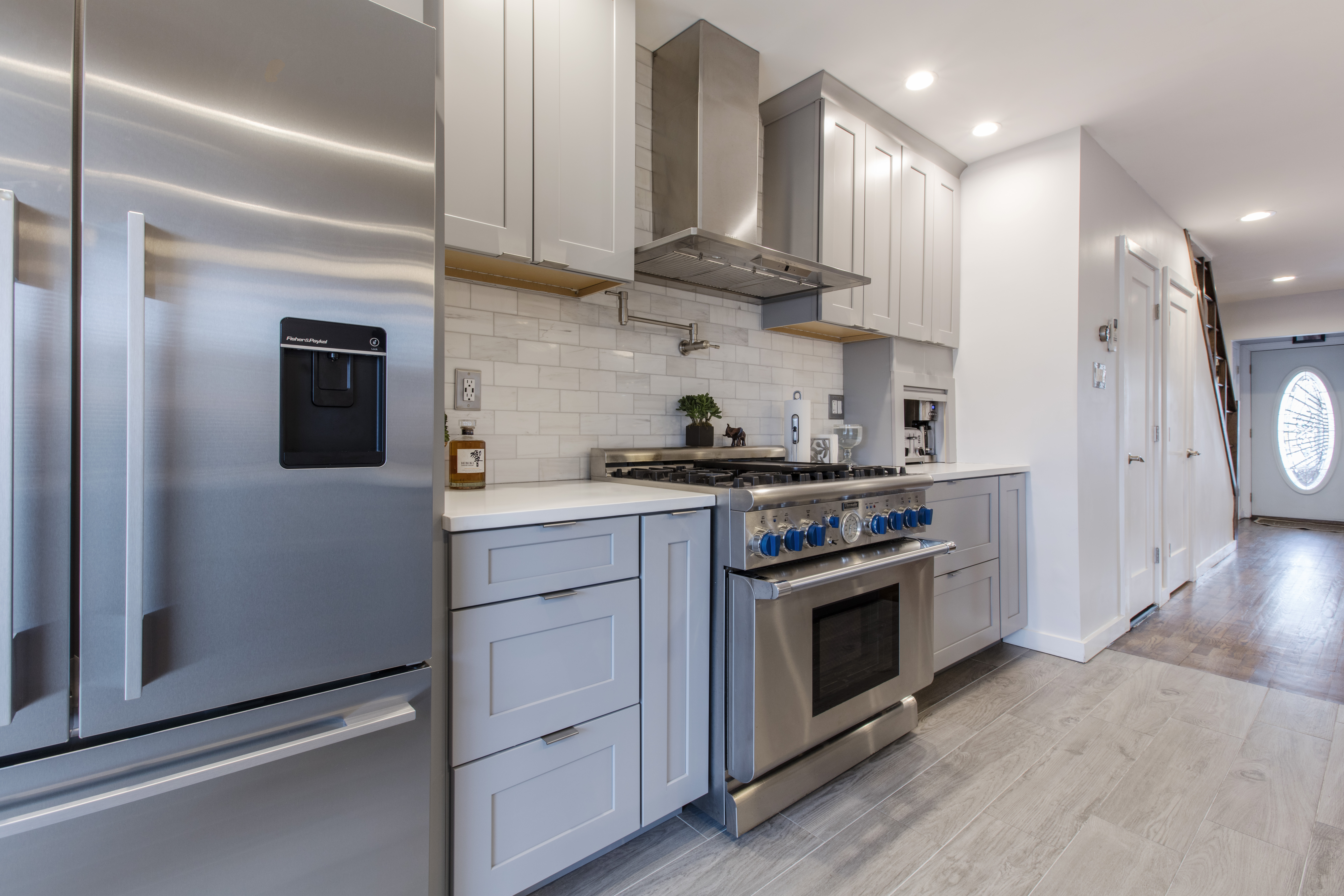 Close-up of a kitchen with gray cabinets, a gas stove, and a stainless steel refrigerator, featuring subway tile backsplash.