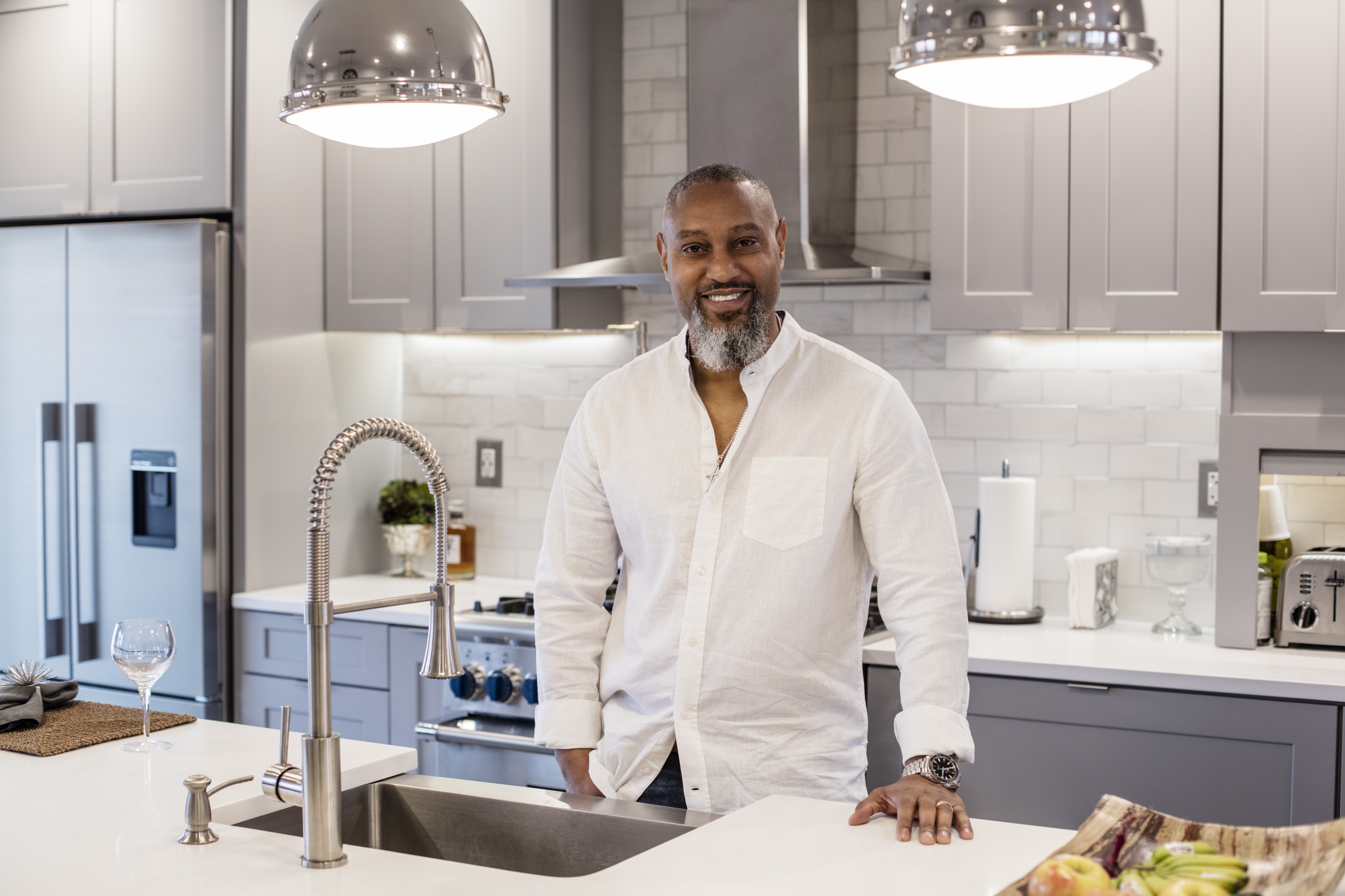 A man stands at a kitchen island with a white countertop, pendant lighting, and stainless steel appliances in the background.