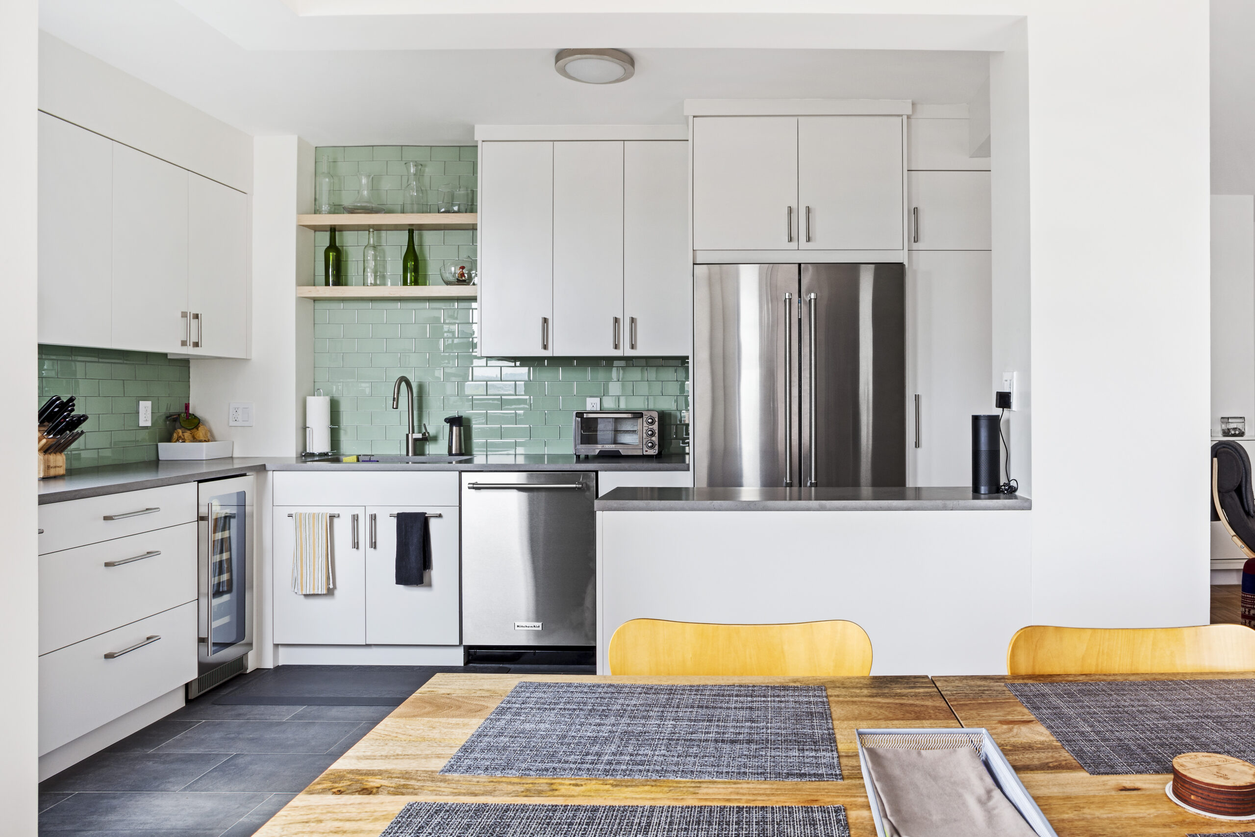 Light-filled modern kitchen with white cabinets, green subway tile, and open shelves, featuring stainless steel appliances.