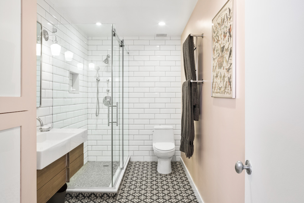 Bright bathroom with glass shower, white subway tile, patterned black-and-white floor, wall-hung wood vanity, pink accent wall, towel bars, and toilet.