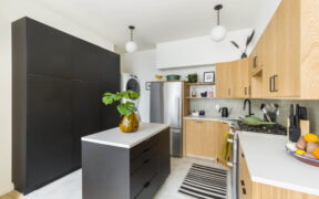 Small kitchen with black island, tall black pantry wall, oak cabinets, and a stacked washer-dryer beside a stainless steel fridge.