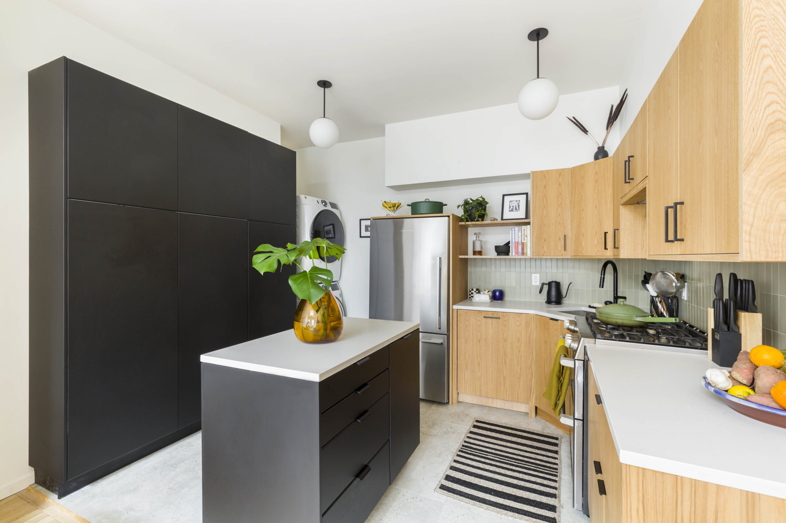 Small kitchen with black island, tall black pantry wall, oak cabinets, and a stacked washer-dryer beside a stainless steel fridge.